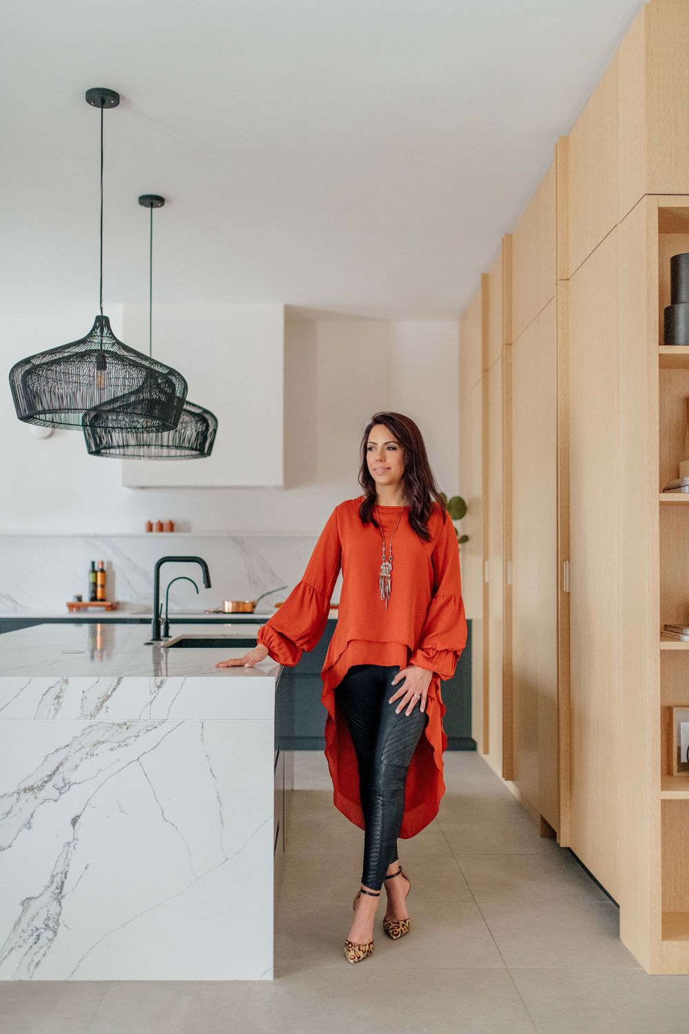 Woman in red blouse leaning on marble kitchen island with black pendant lights and wooden cabinetry.