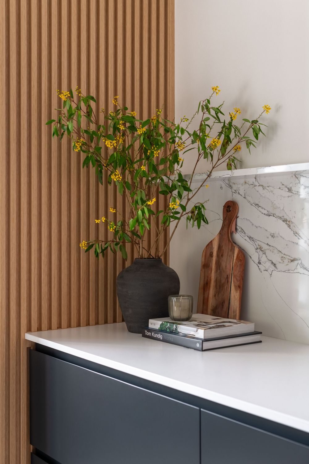Modern kitchen corner with dark vase, yellow flowers, wooden cutting board, and marble backsplash on white countertop.