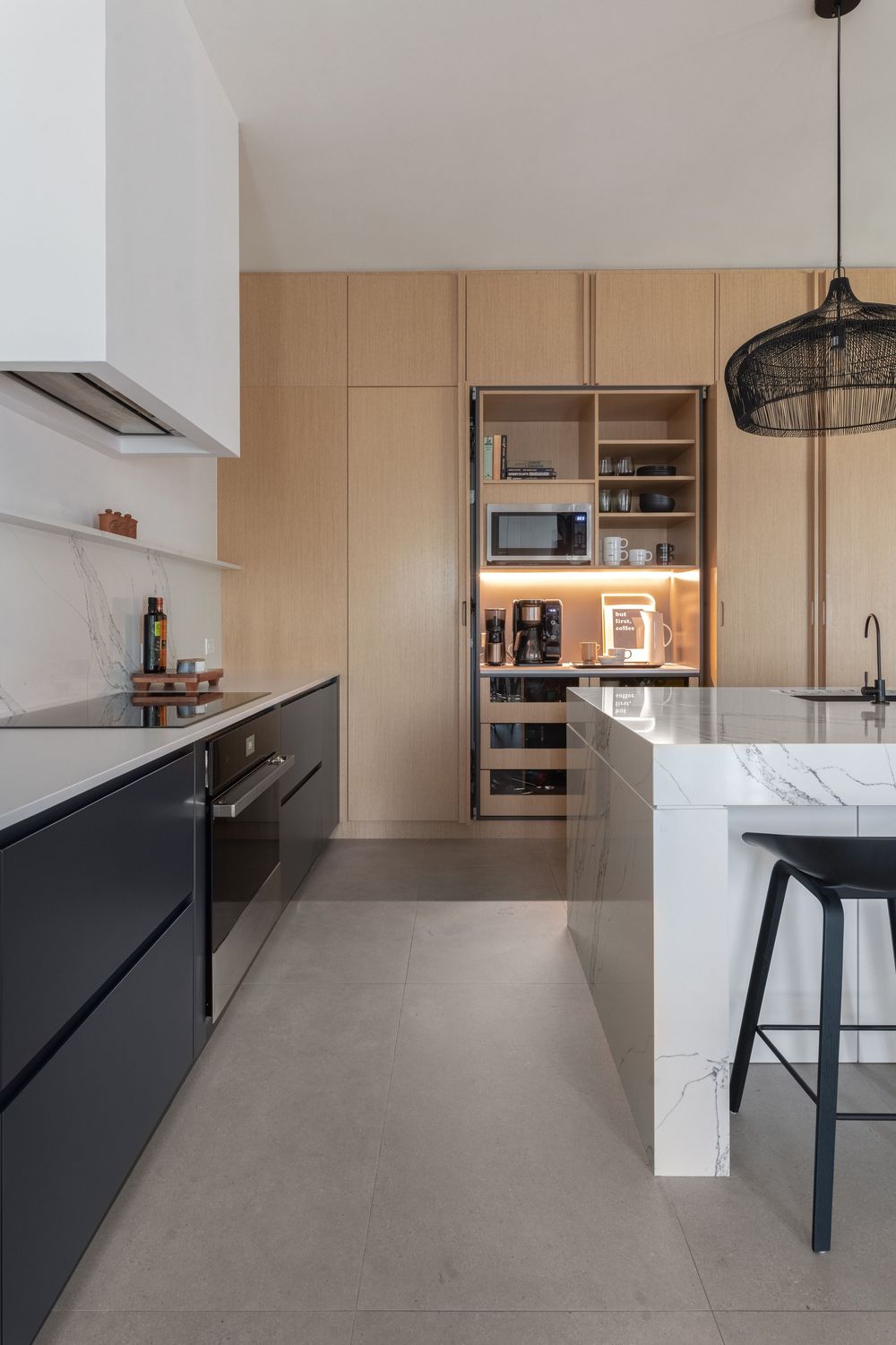 Modern kitchen with marble island, black cabinets, wood storage wall, and pendant light.