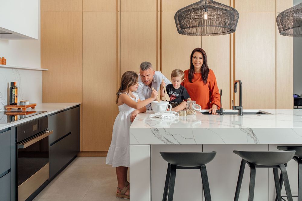 Family of four gathered around a marble kitchen island in a modern home with warm wood cabinetry and sleek appliances.