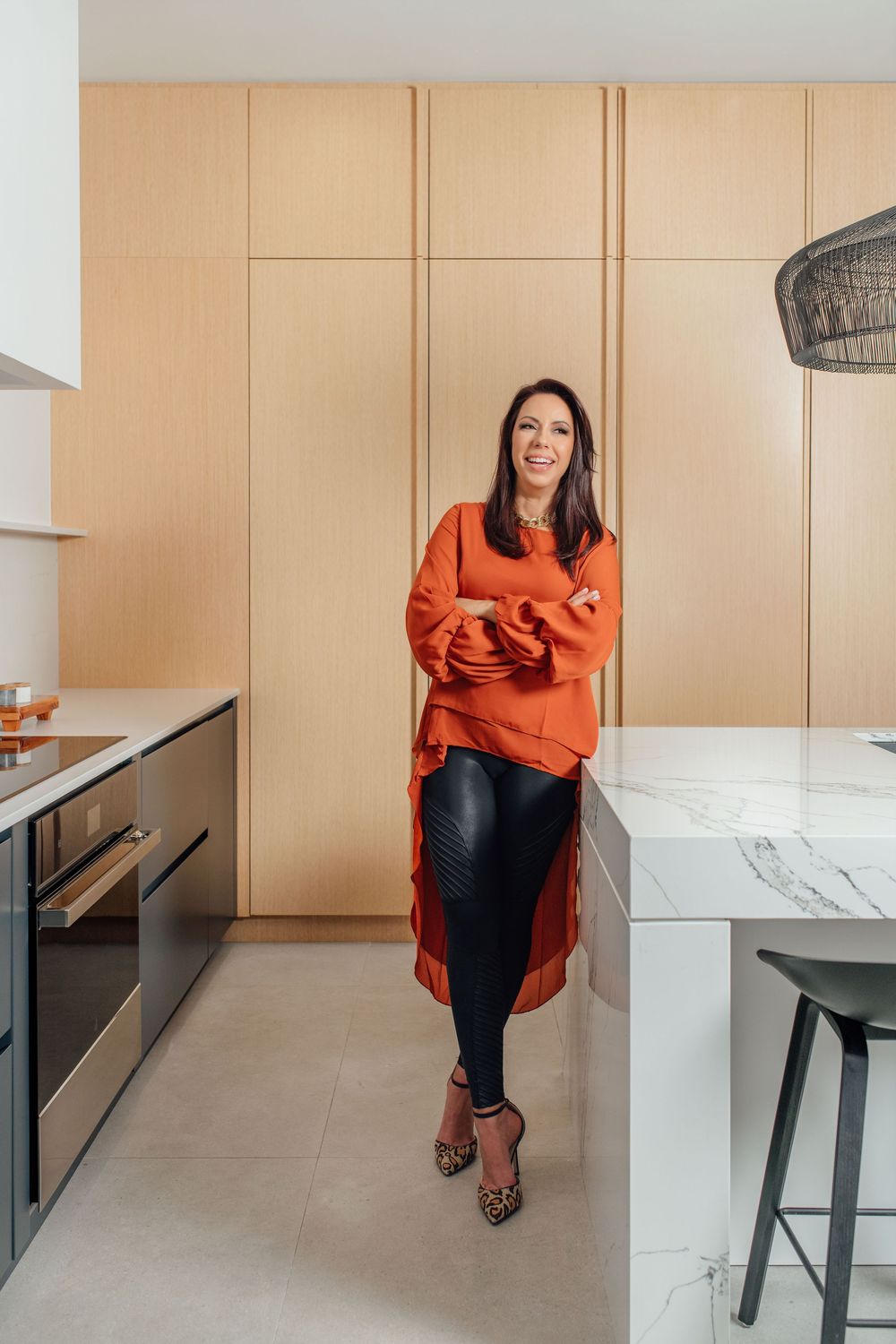 Woman in orange top and black leather leggings poses confidently in a modern kitchen with marble island and wood cabinetry.