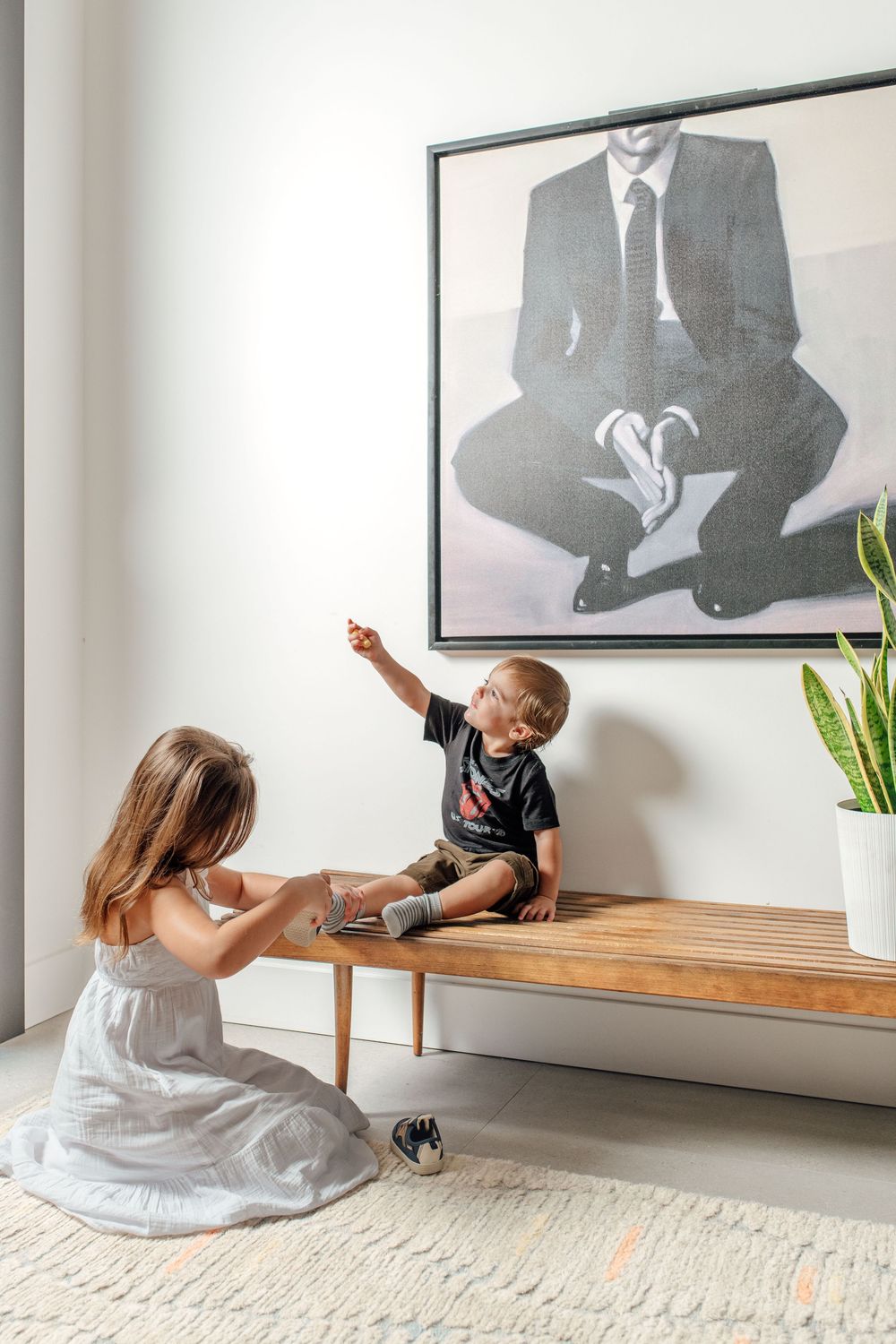 Two children sit on a wooden bench beneath a large framed portrait painting in a modern white interior.