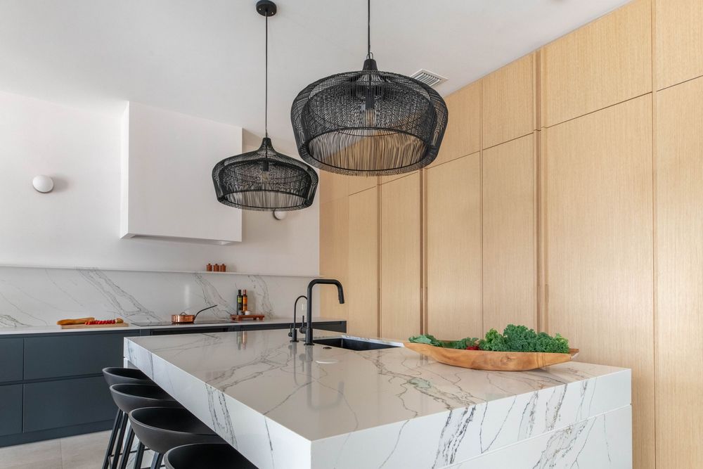 Modern kitchen with marble island, black pendant lights, wood cabinets, and dark bar stools in a bright, elegant space.