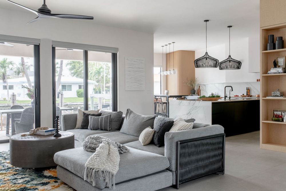 Modern open-plan living room with gray sectional sofa, pendant lights, and kitchen in neutral tones.