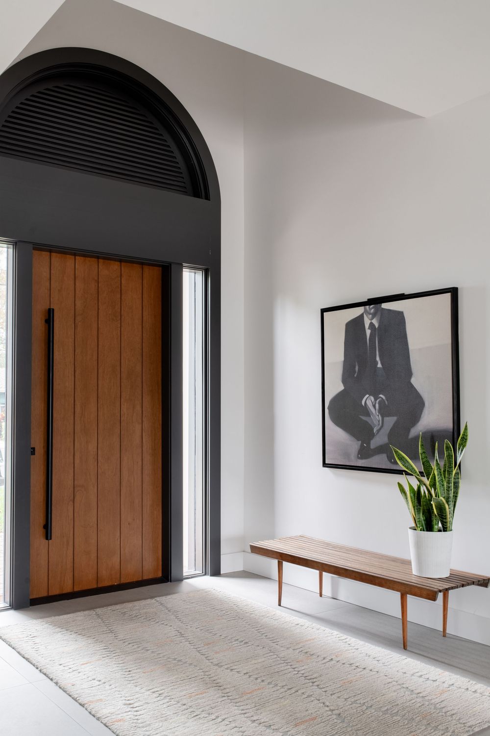 Modern entryway with arched wooden door, mid-century bench, snake plant, and black-framed portrait on white wall.