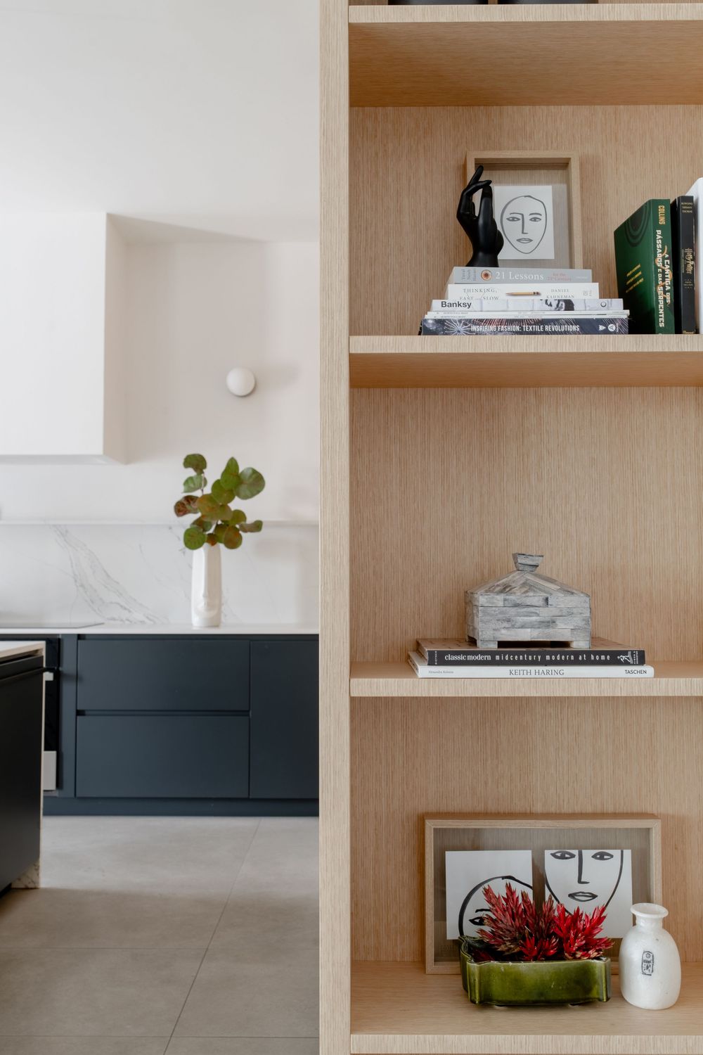Light wood built-in shelving unit displaying books, decorative objects, and face artwork, beside a navy kitchen with marble backsplash.