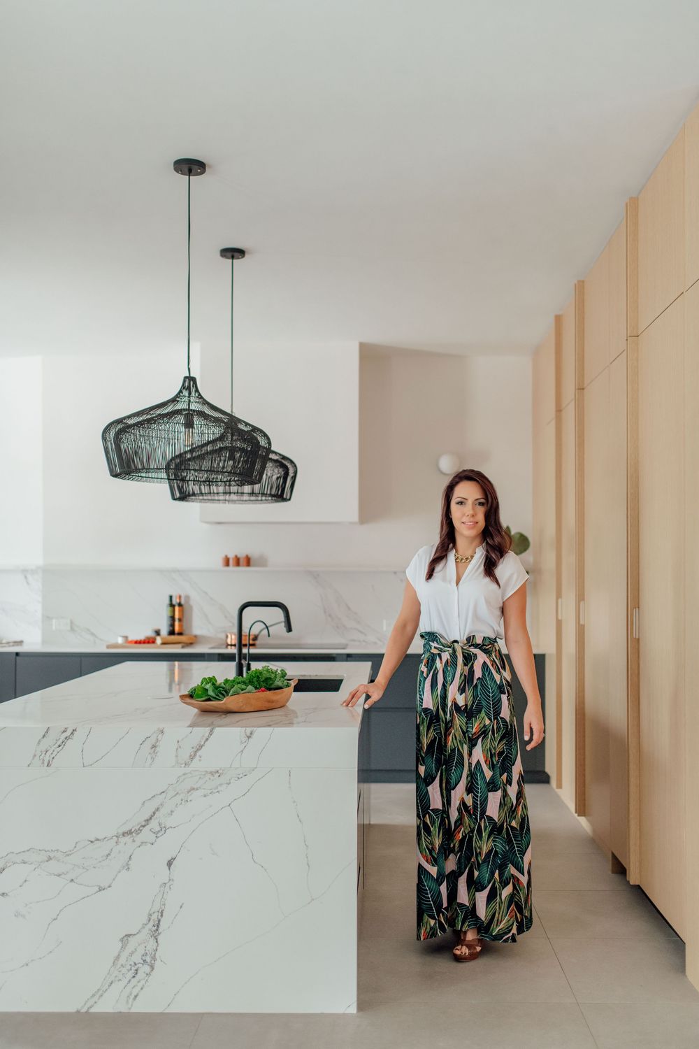 Woman standing beside marble kitchen island with dark pendant lights and timber cabinetry in modern white kitchen.