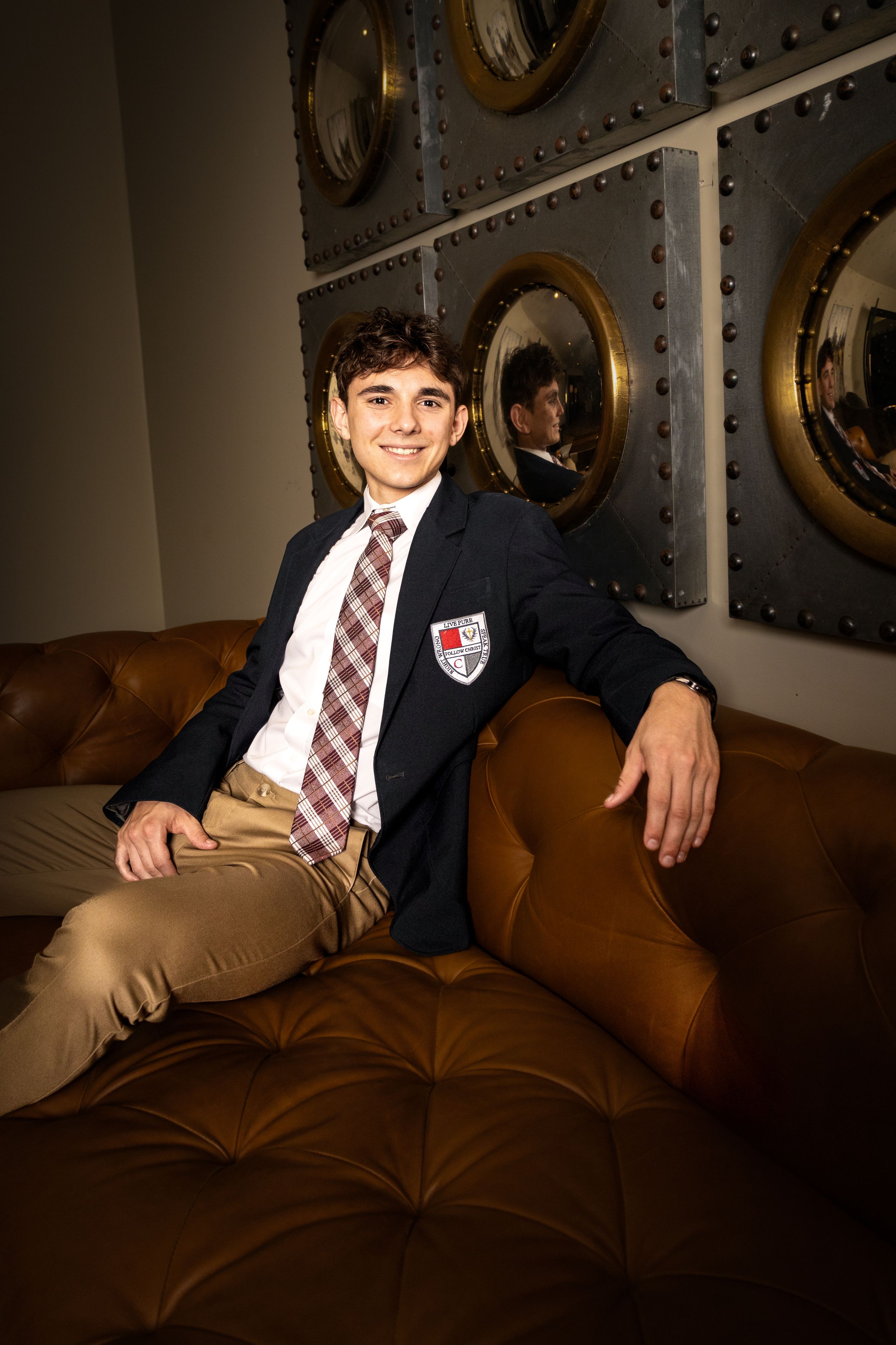 Young man in school blazer with crest, plaid tie, and khakis sits smiling on brown leather sofa near round mirrors.