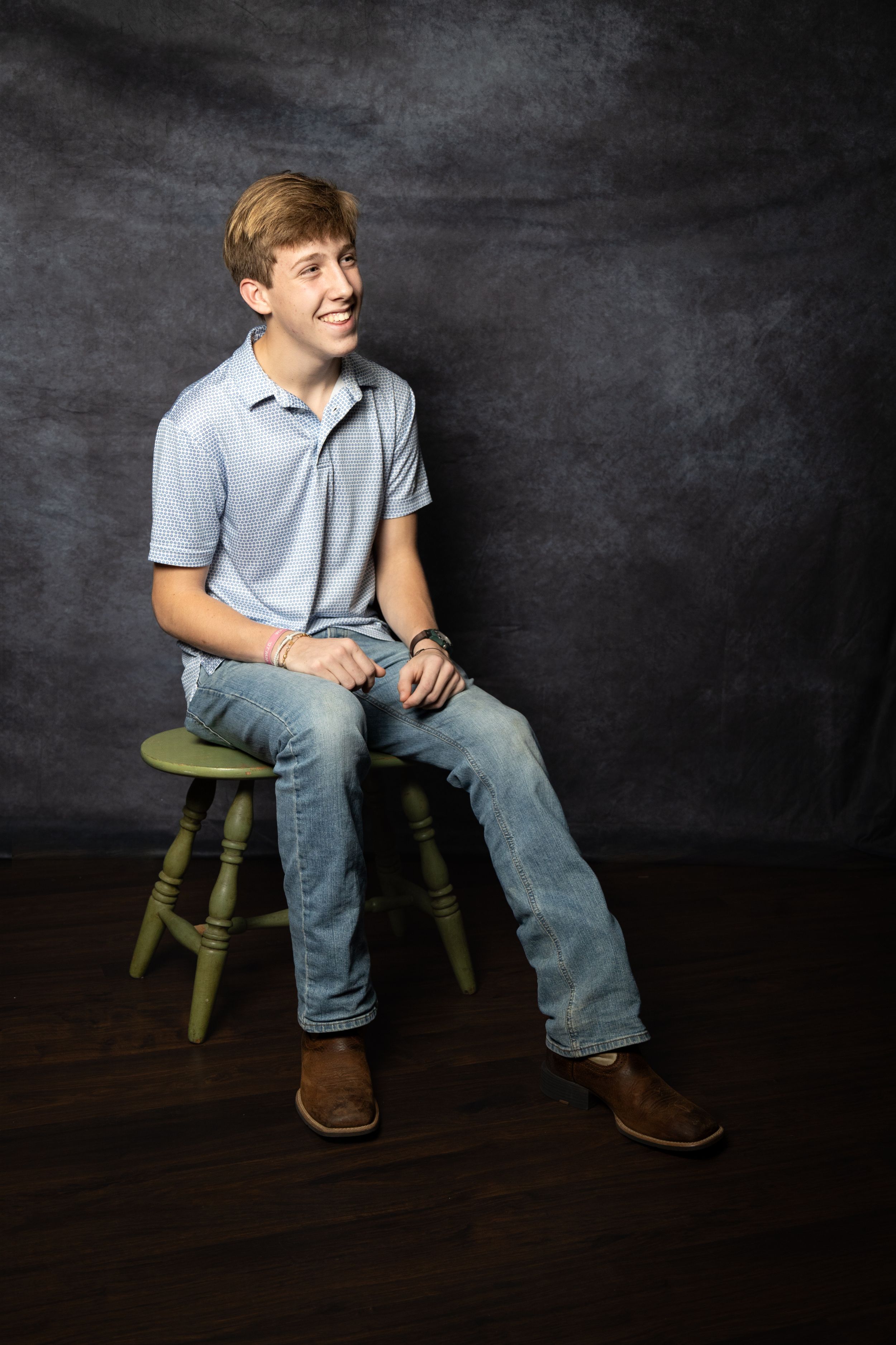 Smiling teenage boy sitting on a green stool in a studio, wearing a blue polo shirt and jeans.