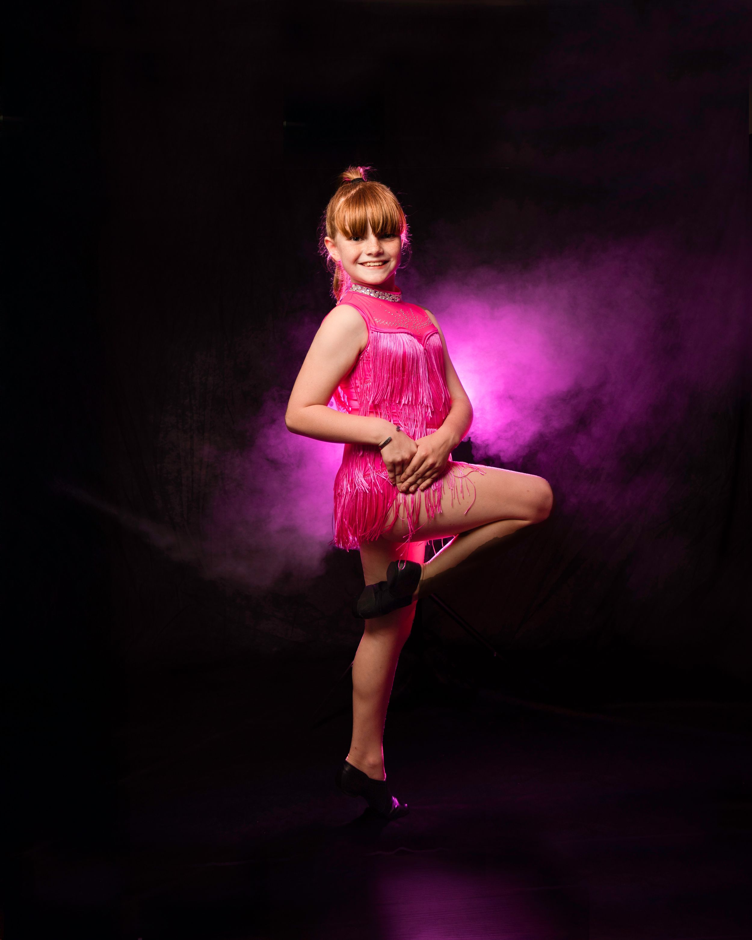 Young dancer in pink fringe costume poses gracefully on stage with dramatic purple lighting backdrop.