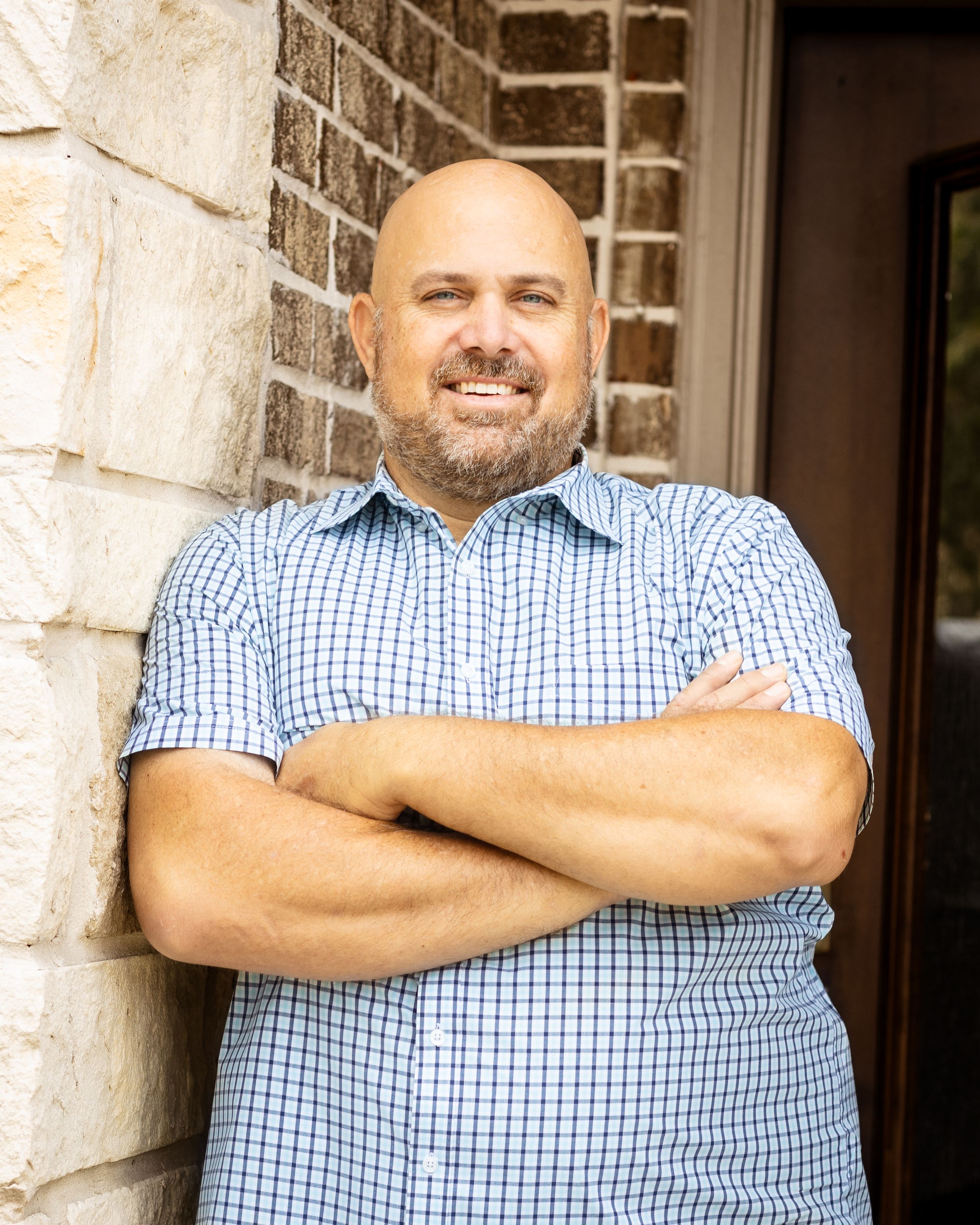 Smiling bald man with beard, arms crossed, leaning against stone wall in blue checkered shirt.