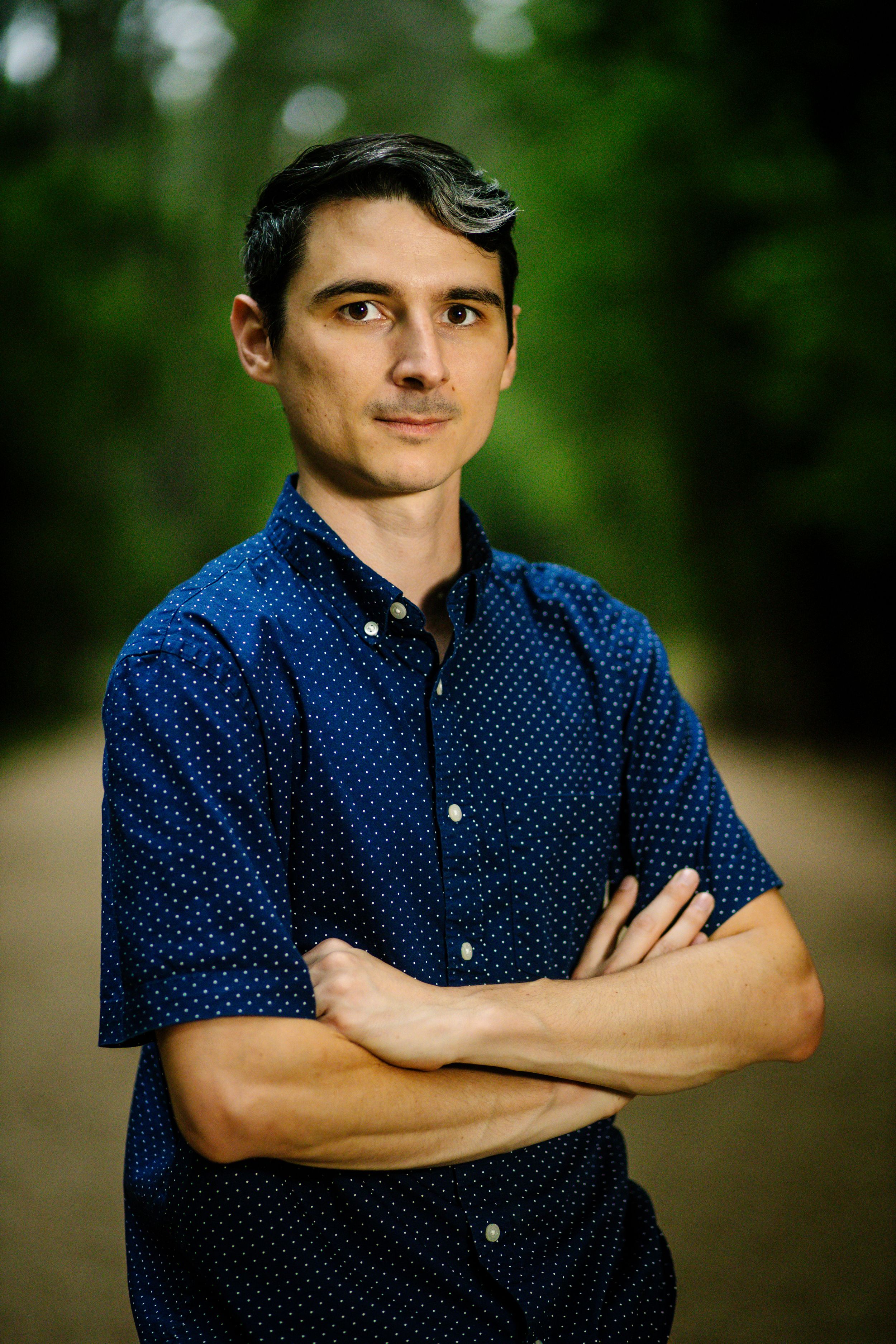 Young man with dark hair, arms crossed, wearing a blue polka dot shirt, standing outdoors with green trees background.