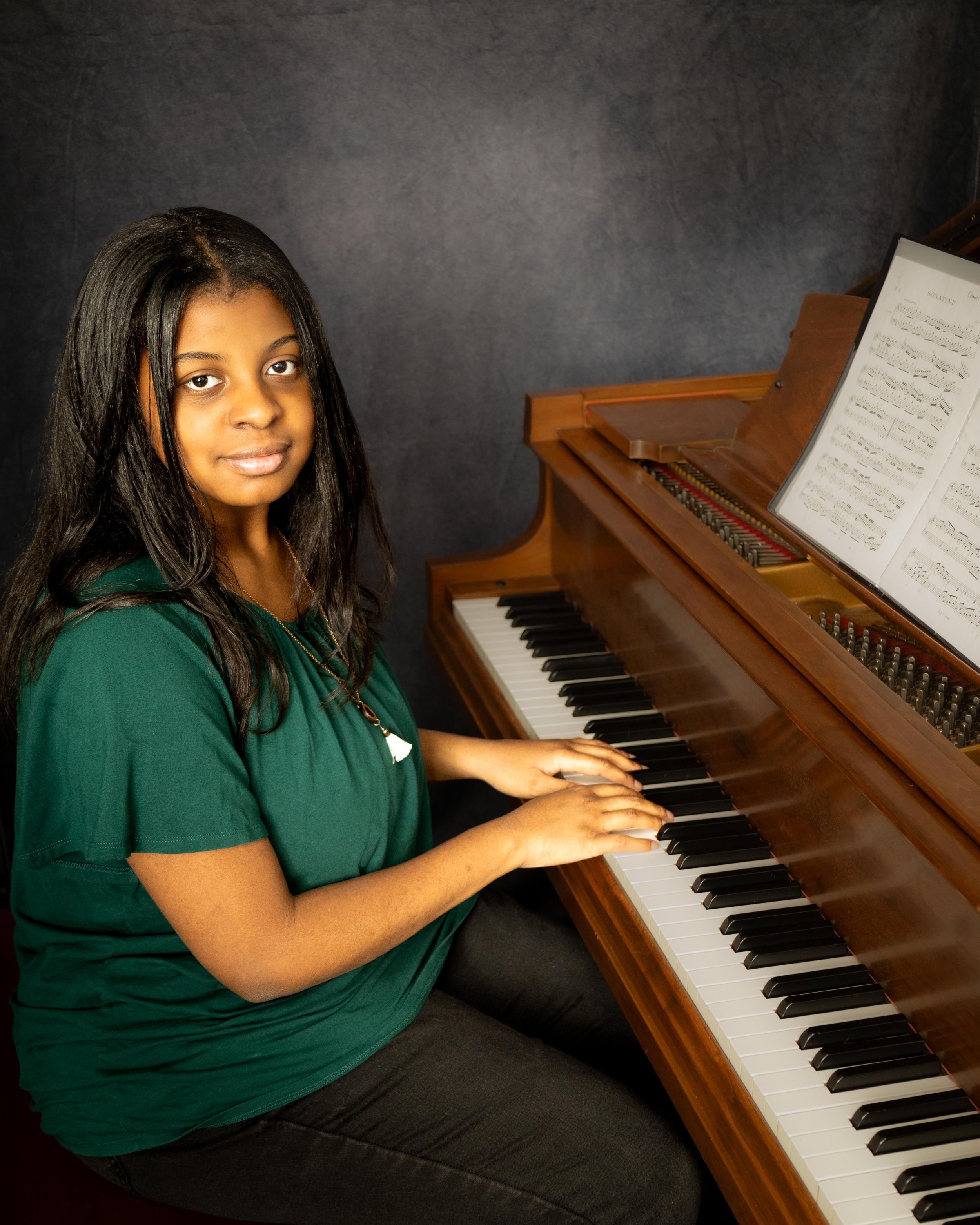 Young woman in green shirt posing at a wooden grand piano with sheet music, smiling at camera.