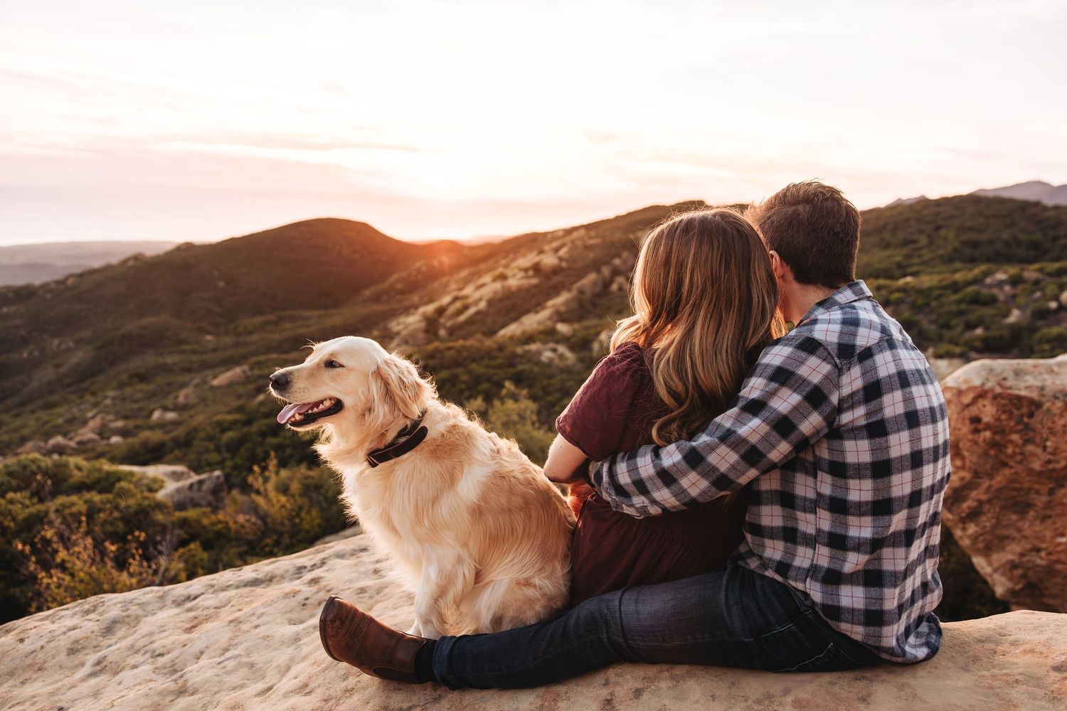 Couple sitting on a rocky hilltop with their golden retriever dog, enjoying a scenic mountain sunset view.