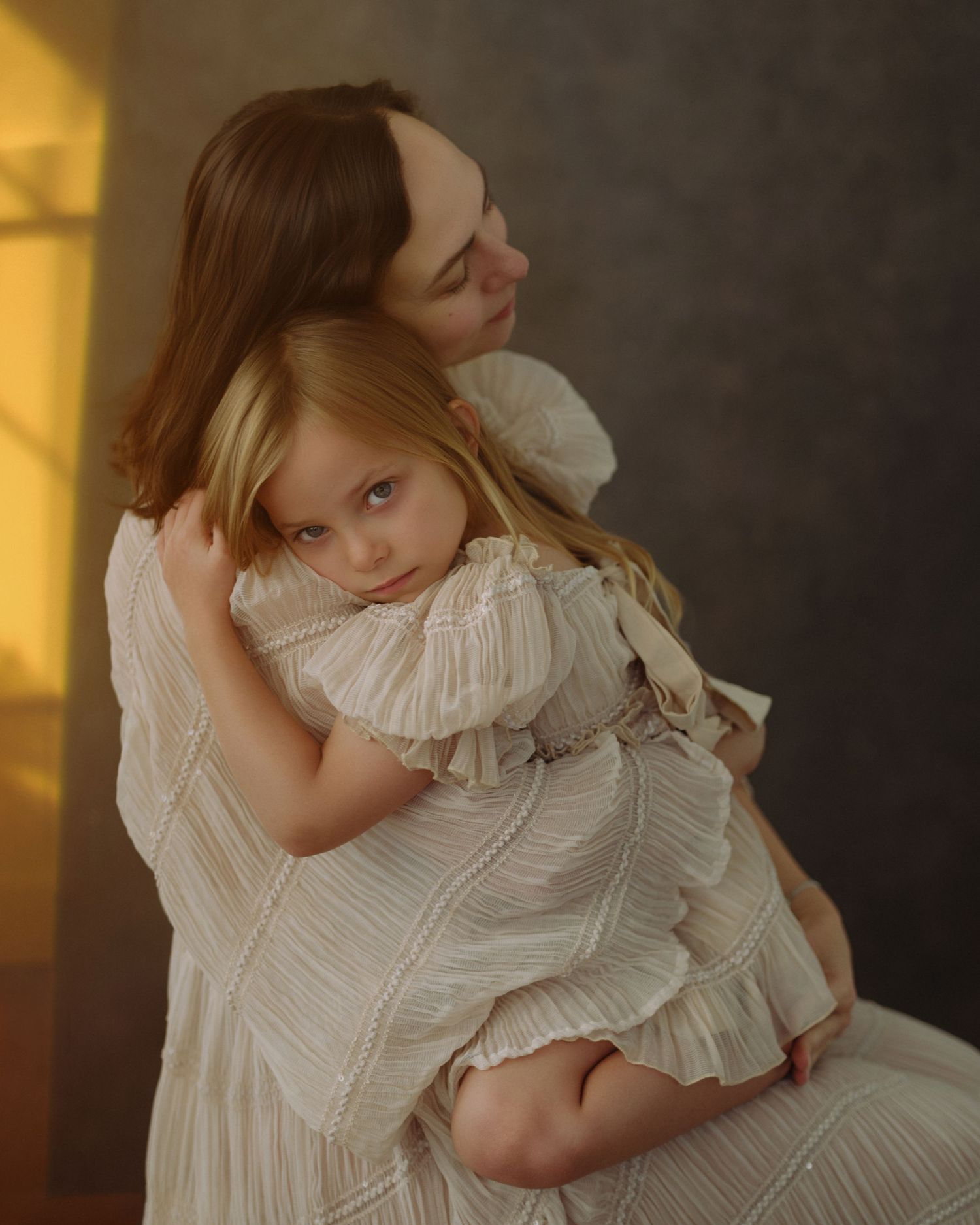 A mother in a cream striped dress holds her young blonde daughter in a matching ruffled dress in a soft, warm-toned portrait.