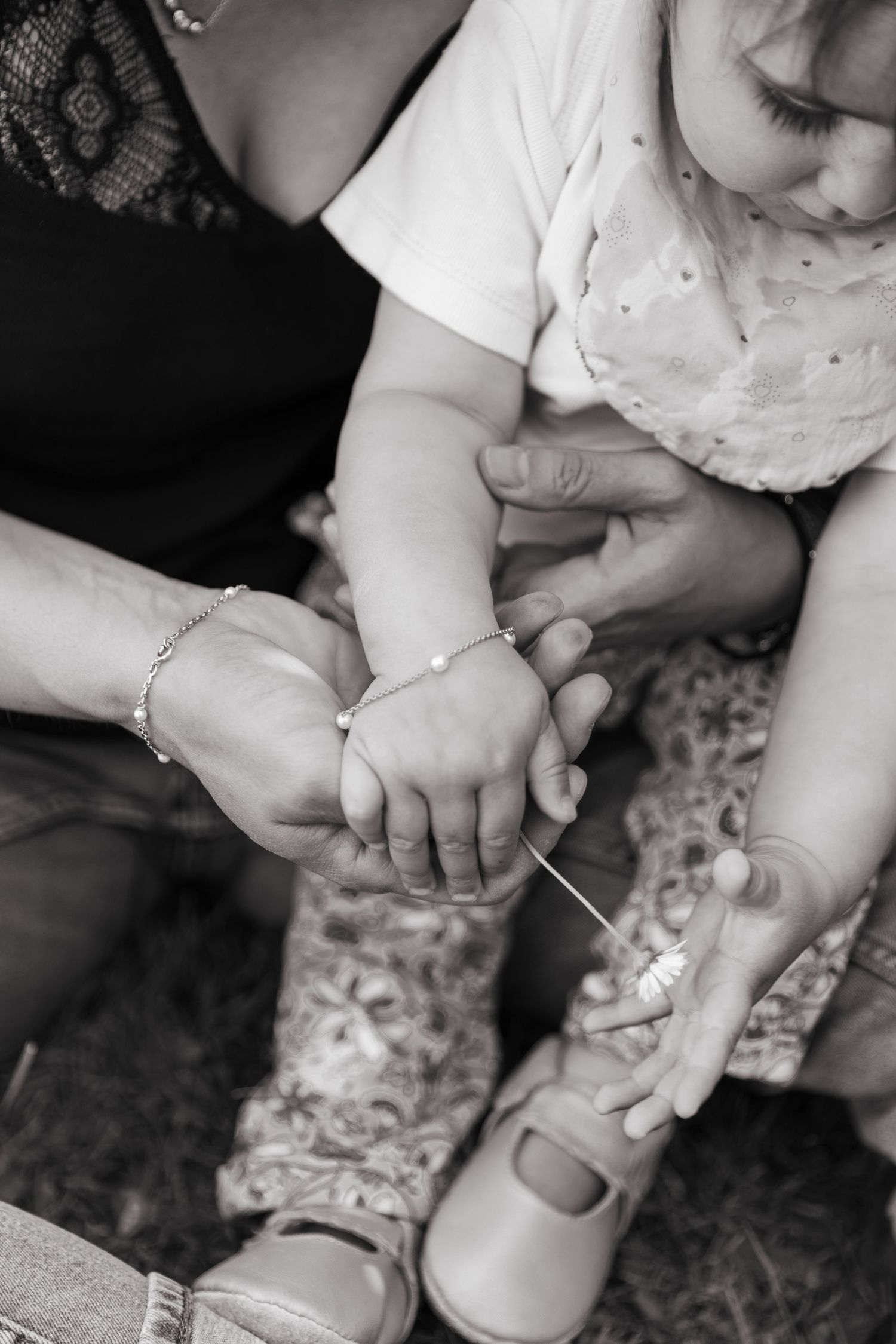 Black and white photo of adult hands helping a toddler girl with a bracelet and shoe.