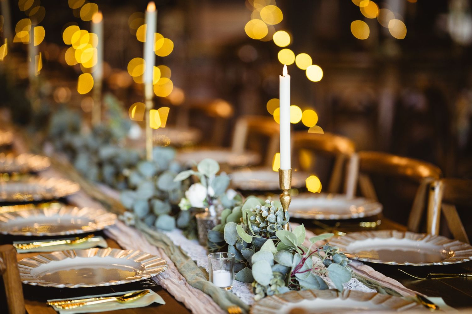 Elegant table setting with glowing candles and eucalyptus garland on rustic wooden surface with golden bokeh lights.