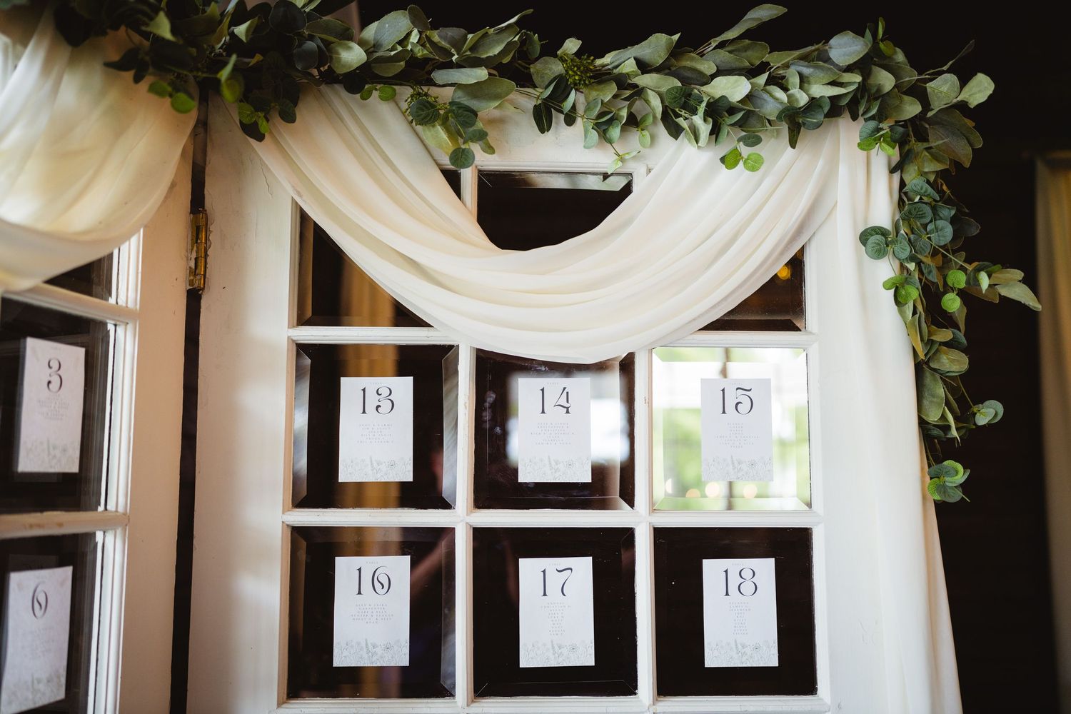 Wedding seating chart displayed on black frames under elegant white draping and greenery garland decor.