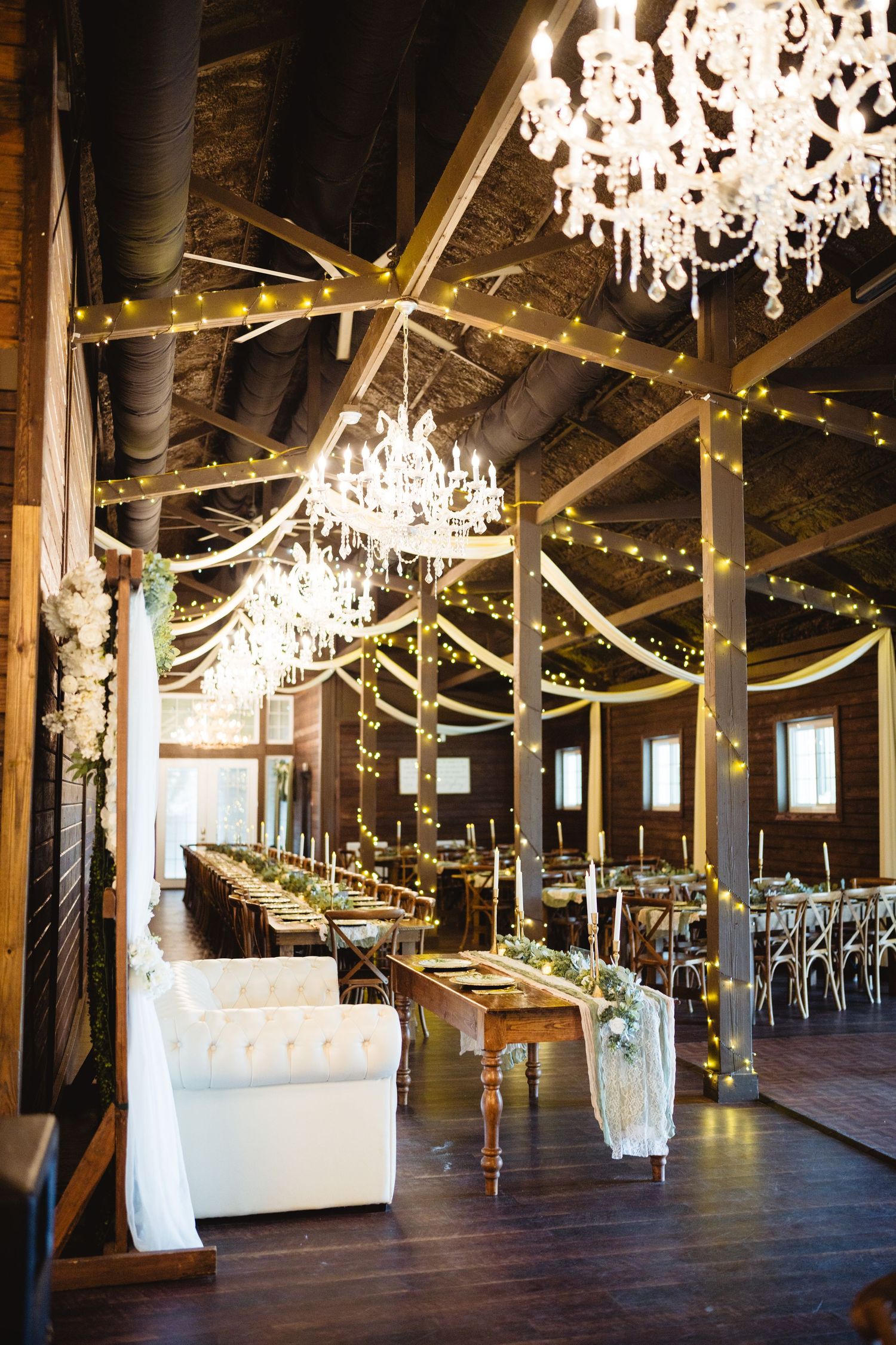 A rustic barn wedding venue with elegant crystal chandeliers, wooden beams, and romantic white draping along the ceiling.