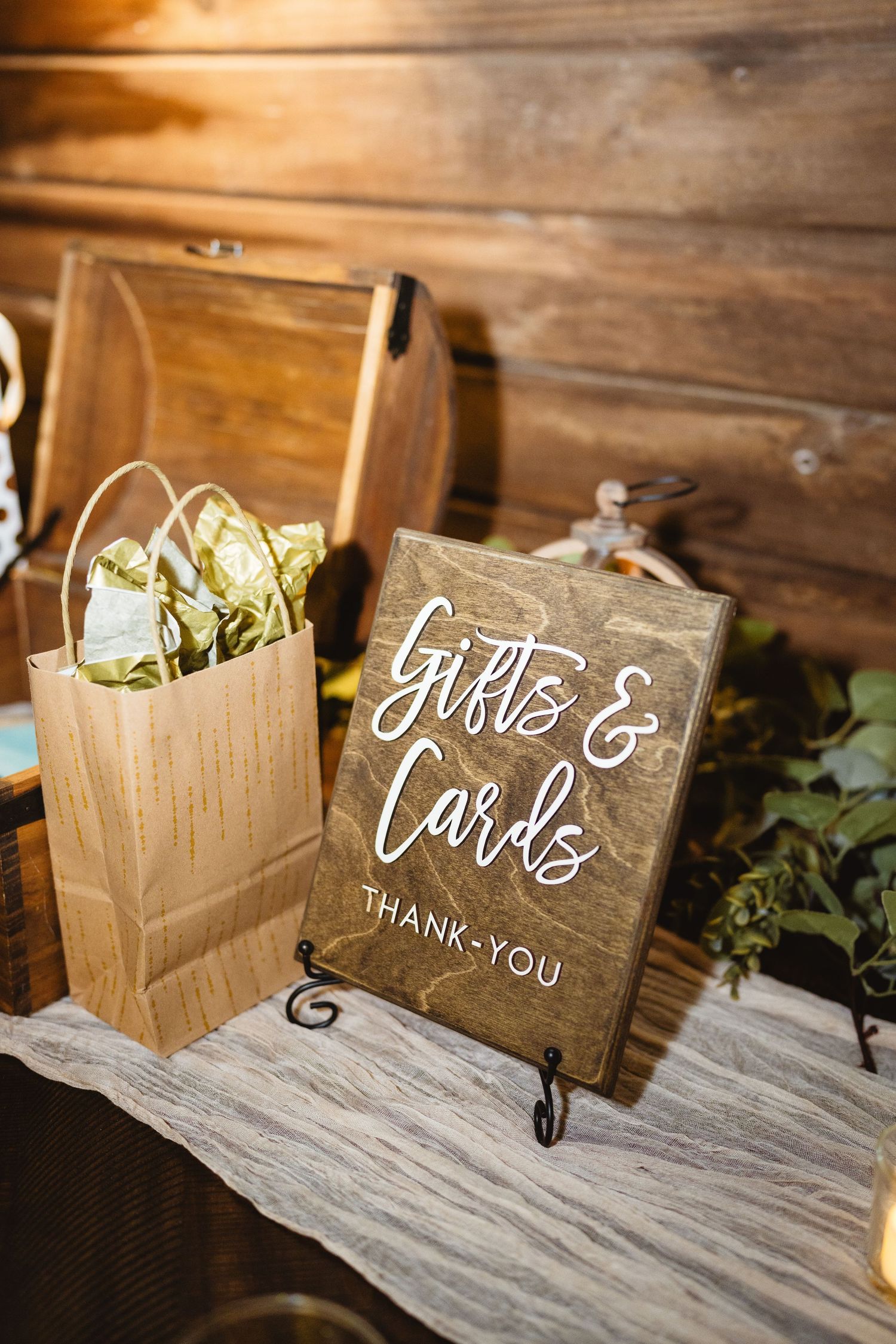 A rustic wooden 'Gifts & Cards' sign displayed on a stand next to a brown paper gift bag at a wedding reception table.