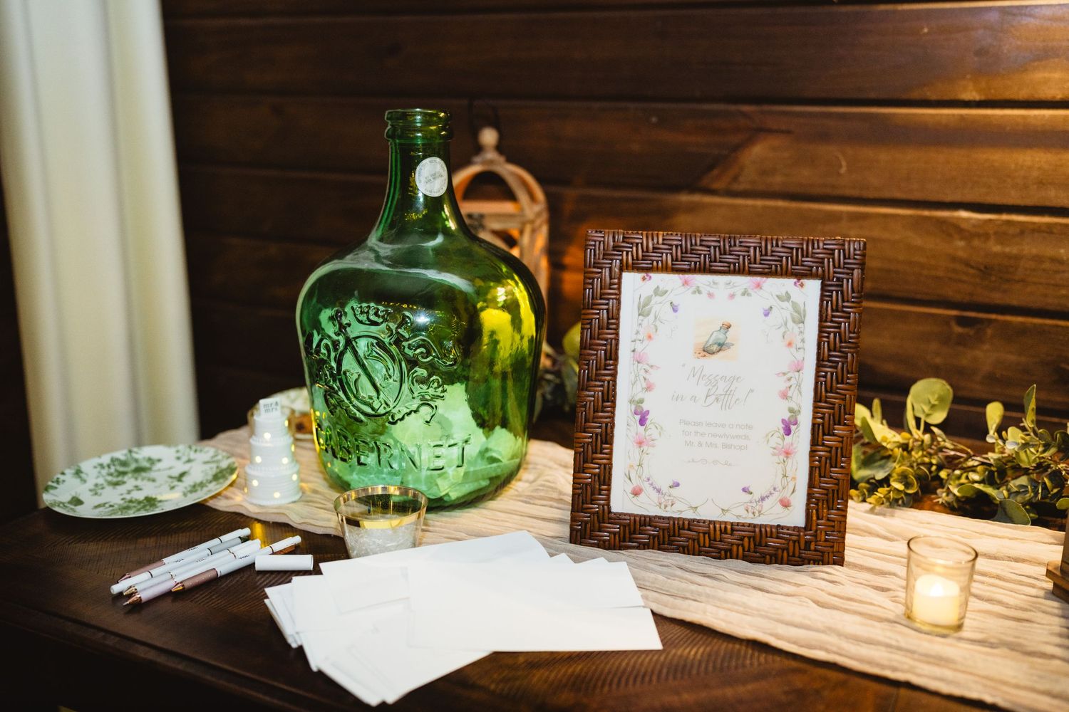 Rustic wedding decor setup featuring vintage green glass bottles and frames on a wooden table with romantic lighting.