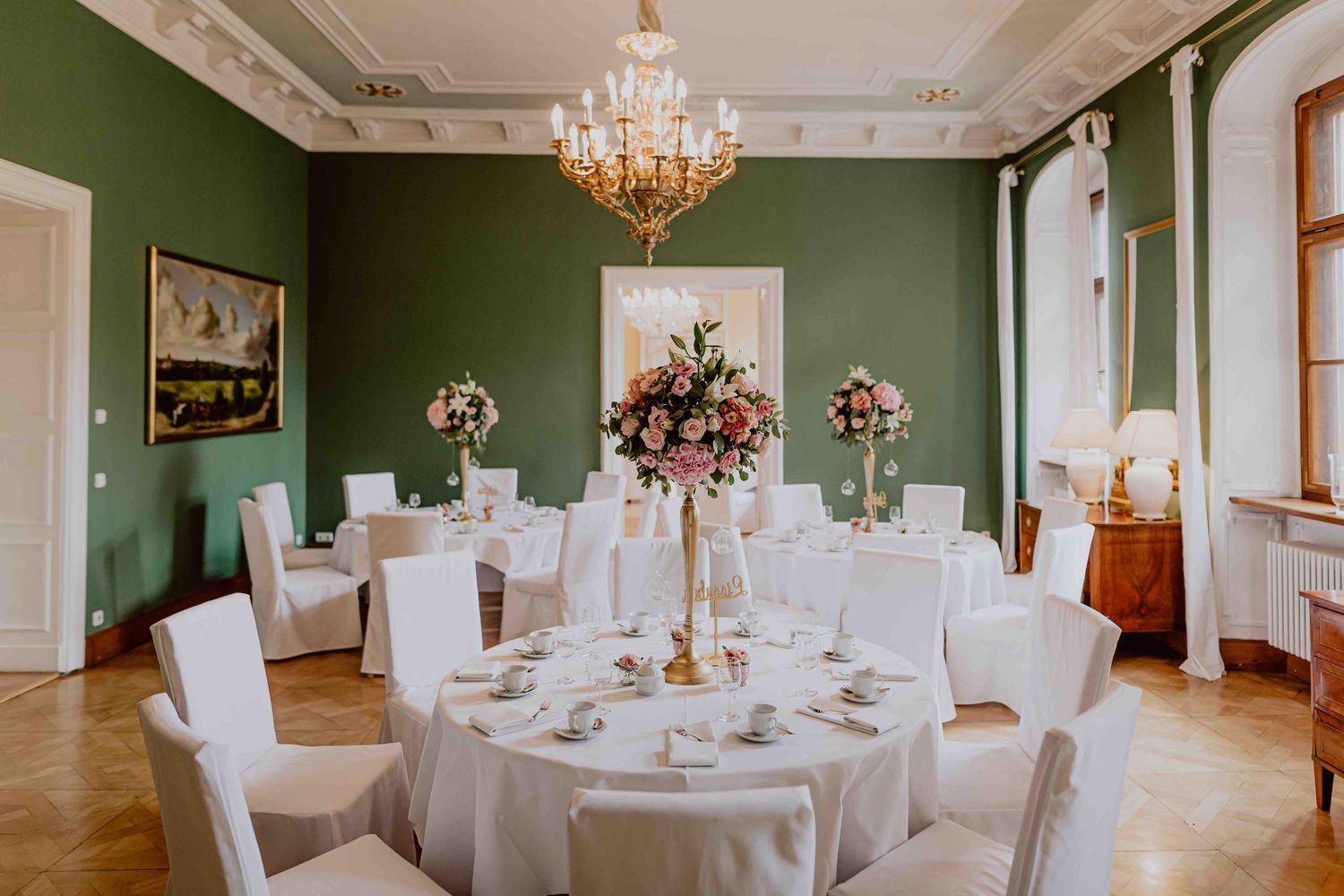 Elegant wedding reception venue with sage green walls, crystal chandelier, white tablecloths and pink floral centerpieces.