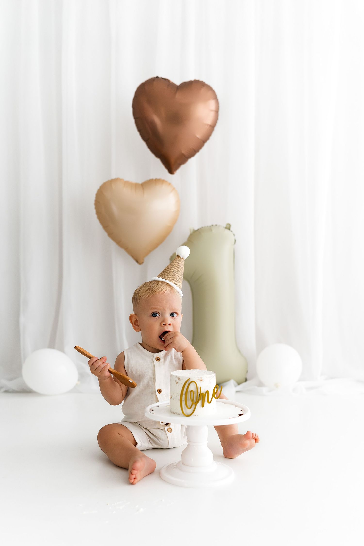 Minimalist portrait of a baby in white with heart balloons and birthday decorations on a light backdrop.