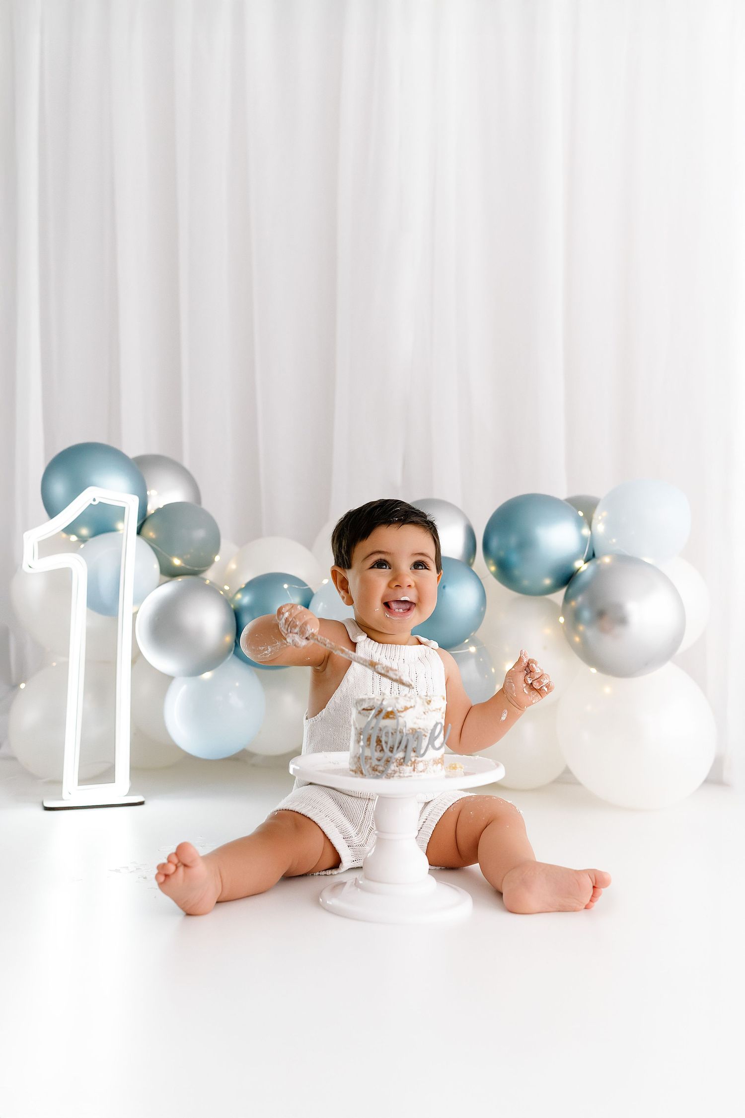 Baby in white onesie poses on white backdrop with silver and blue balloons and number one decoration for first birthday.