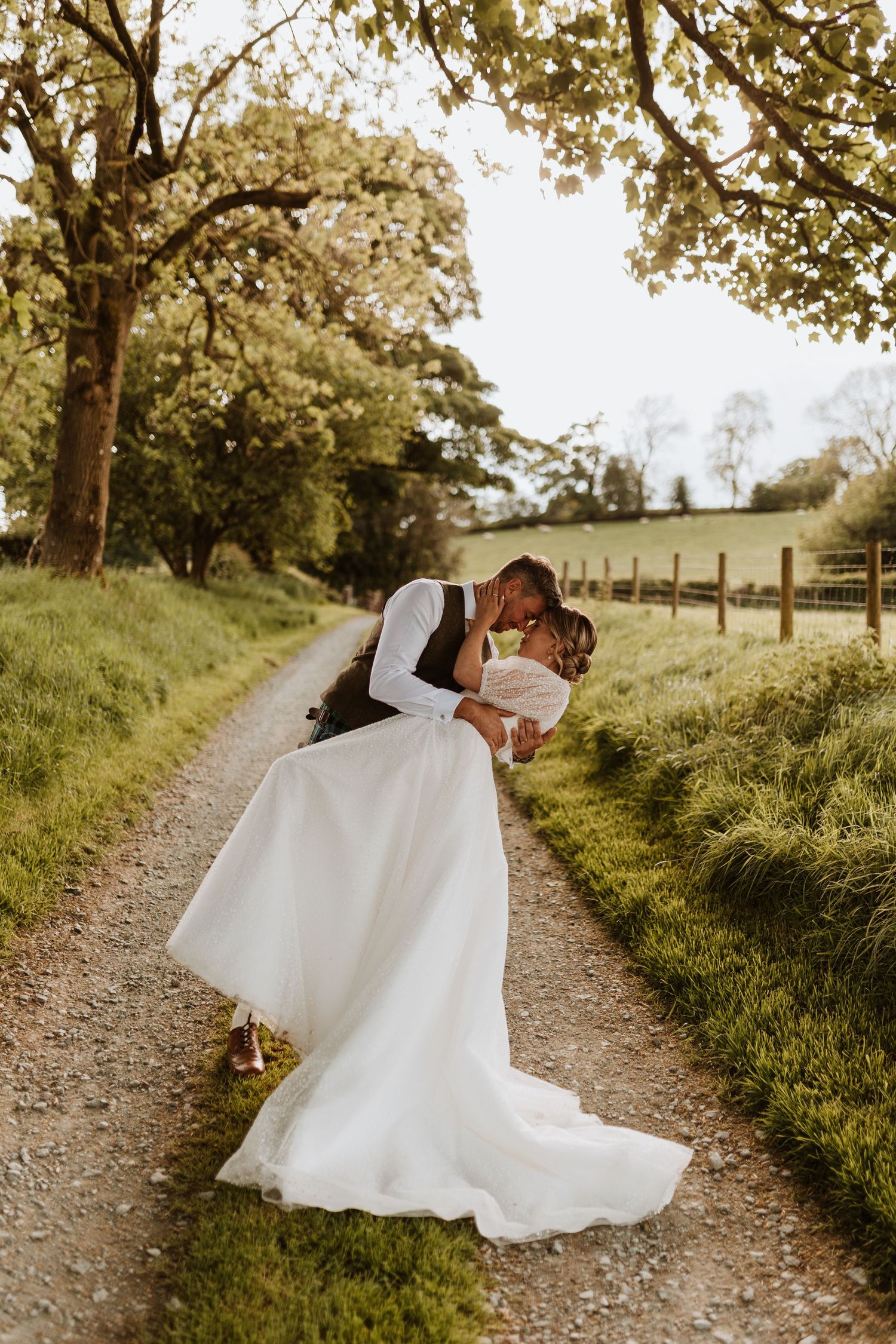 A romantic wedding couple dances on a tree-lined dirt path in autumn, with the bride's white dress flowing gracefully.