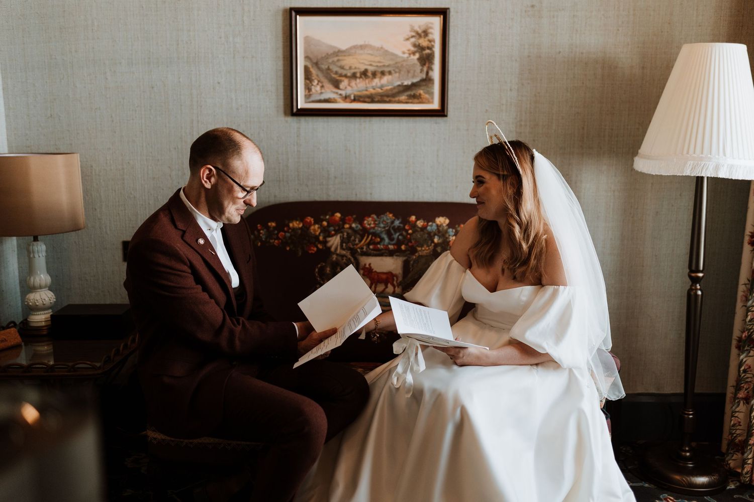 A couple shares a quiet moment in formal attire while reading together on a vintage sofa in warm lighting.