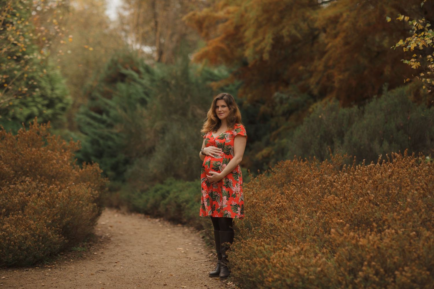 A person in a red floral dress walks along a winding garden path surrounded by autumn foliage and evergreens.