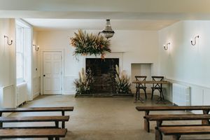 Interior room with wooden benches, stone fireplace, white walls and floral arrangements in minimalist ceremony space at Aswarby Rectory.