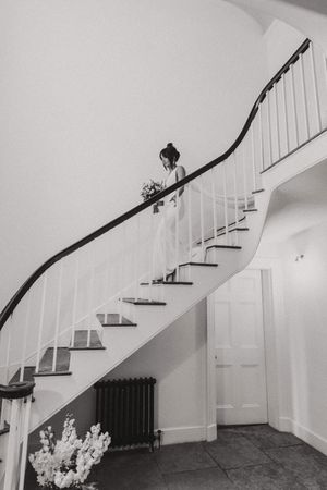 A black and white photograph shows someone in a wedding dress descending an elegant curved staircase with a white railing at Aswarby Rectory