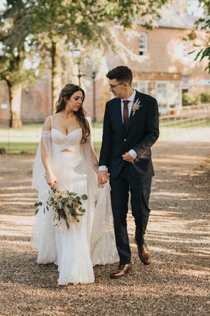 A couple in wedding attire walk together on a leaf-covered path surrounded by autumn trees at Aswarby Rectory.
