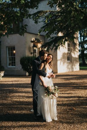 A couple shares a tender moment in evening sunlight outside a white country house surrounded by trees at Aswarby Rectory.