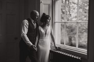 A wedding couple in formal attire stands together by a bright window in an elegant black and white photograph at Aswarby Rectory.
