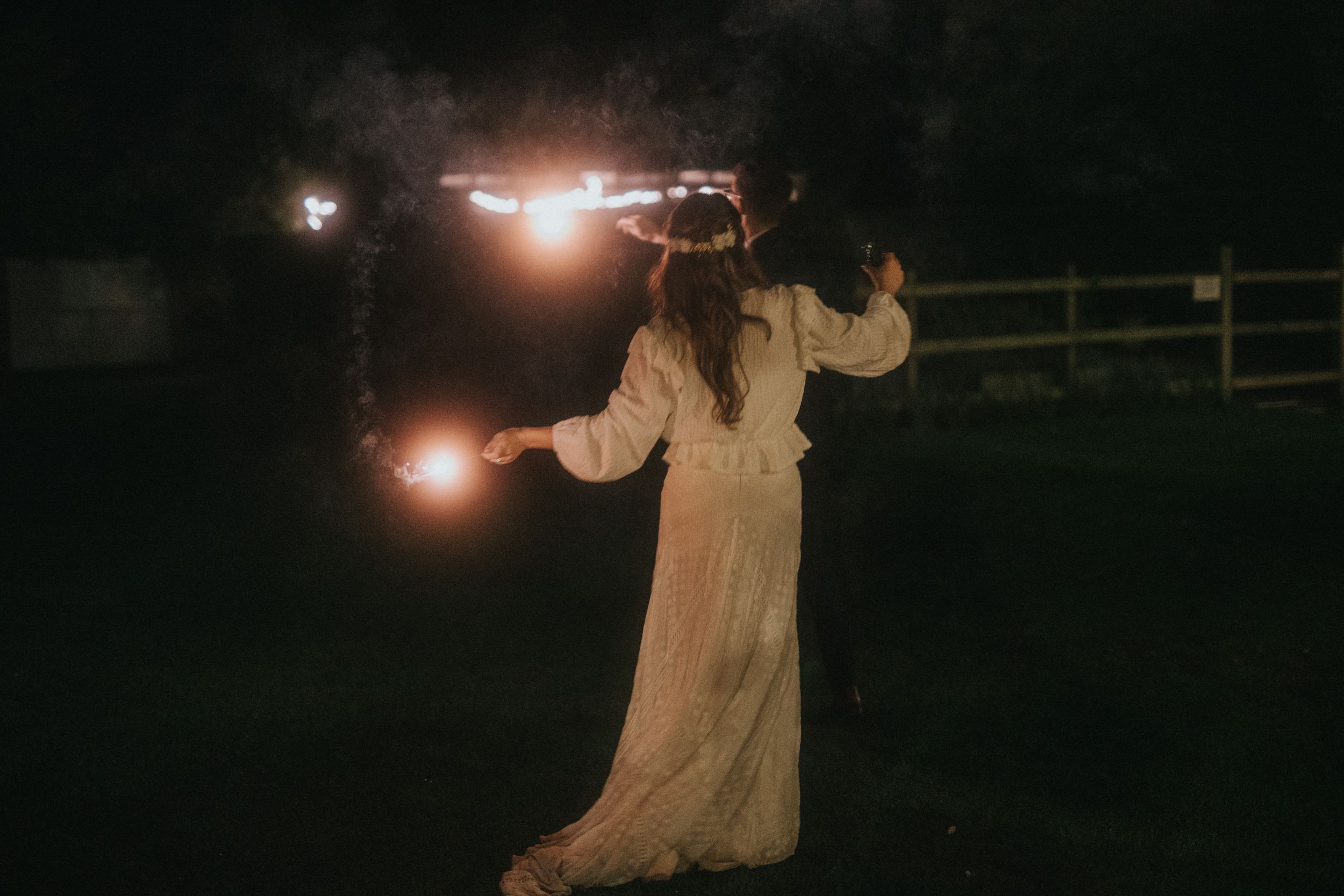 A person in a flowing white dress holds sparklers while standing outside at night near a wooden fence at Aswarby Rectory.