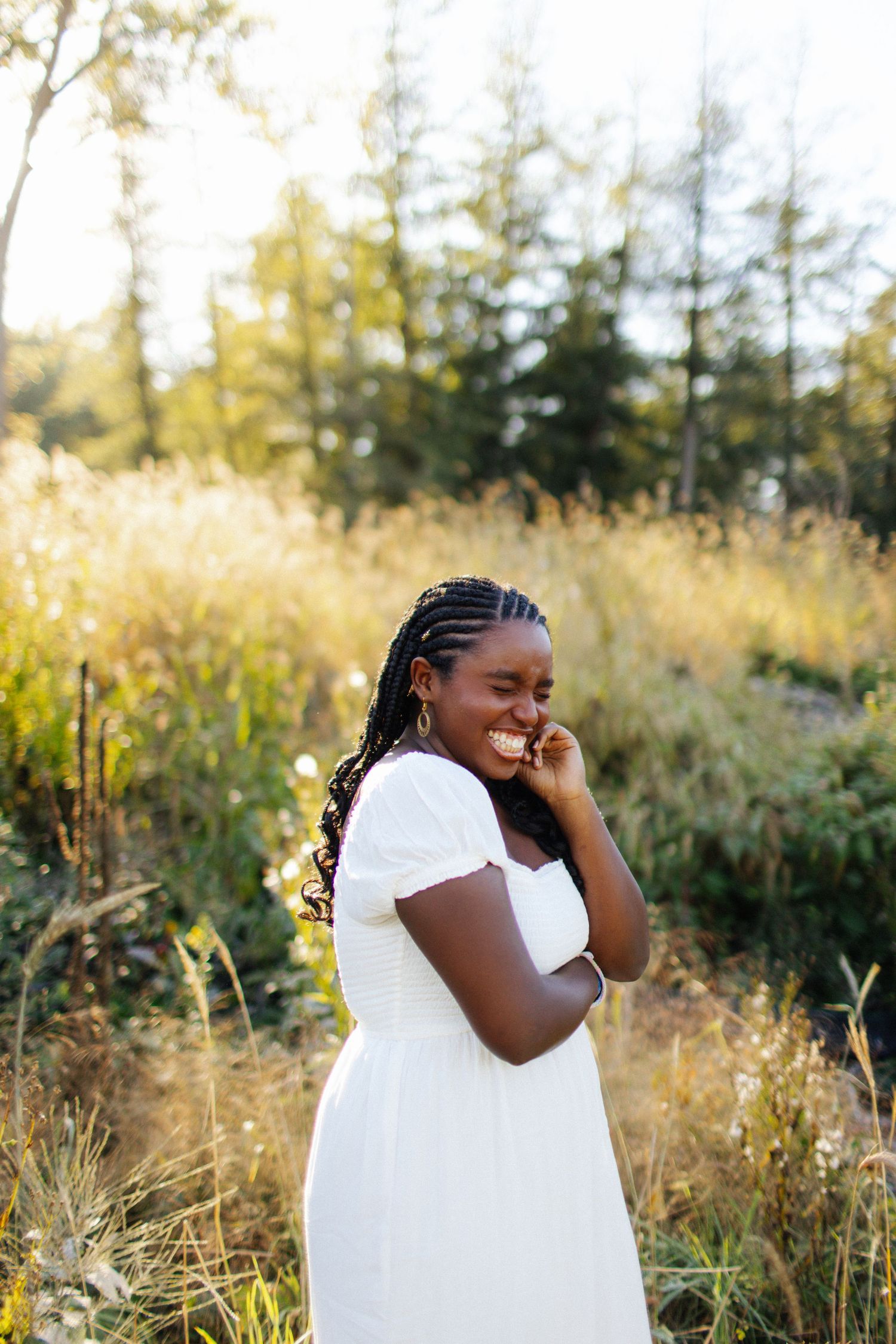 Person in white dress poses in golden field during sunset with evergreen trees in background.