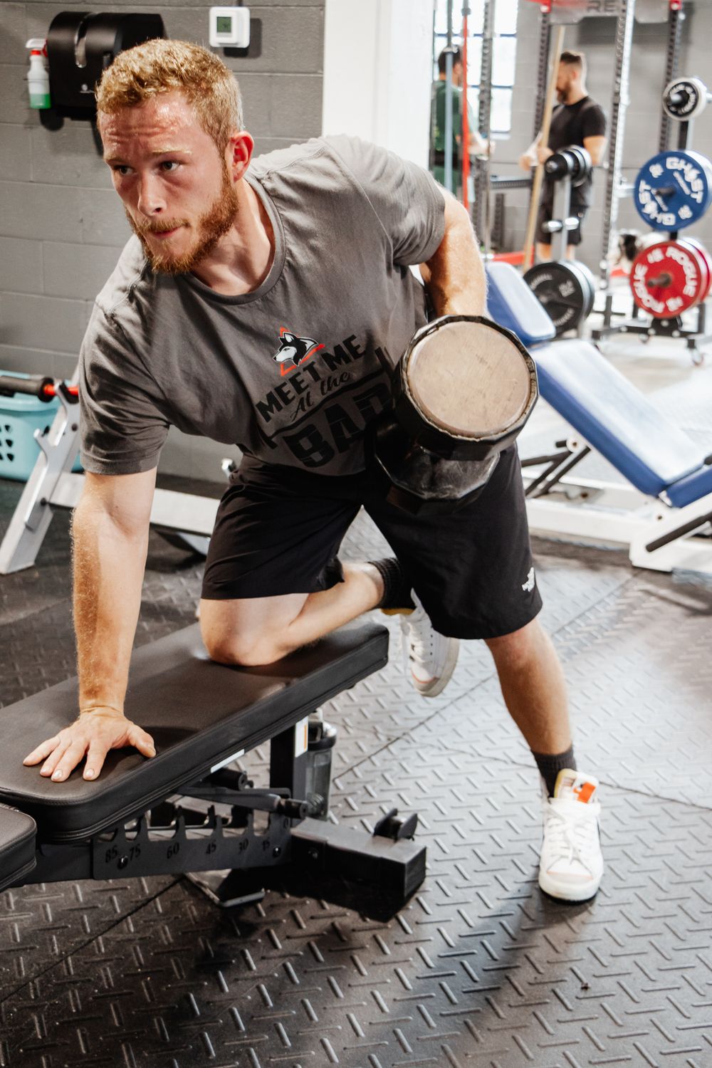 Focused man performs a single-arm dumbbell row on a bench, wearing a "Meet Me at the Bar" gym shirt