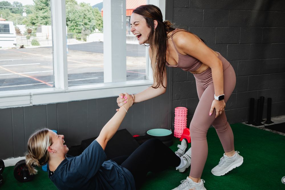 woman in activewear laughs while helping her friend off the gym floor, sharing a joyful and supportive workout moment.