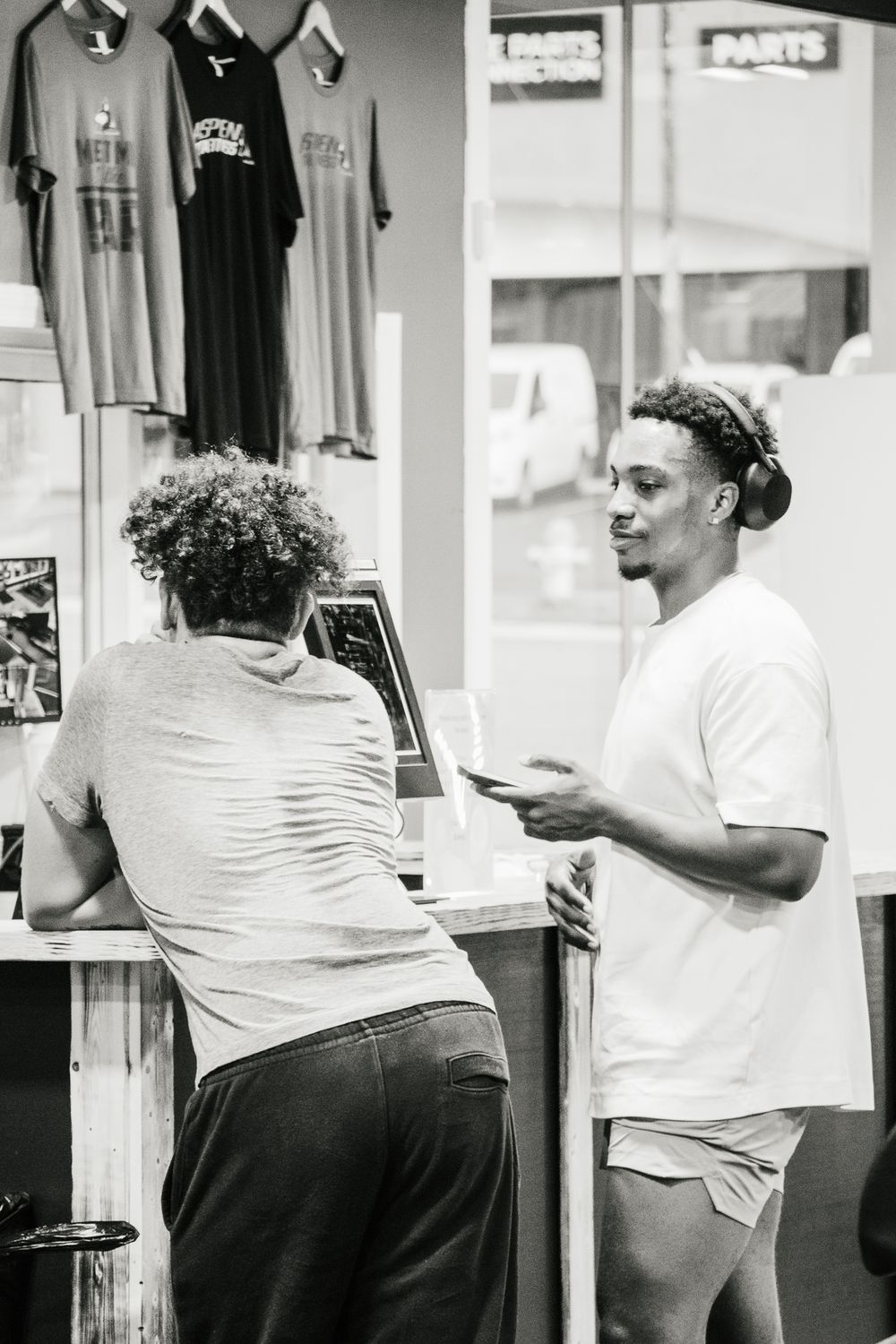 Black and white photo of two men talking at a gym check-in counter, one using a touchscreen and the other holding a phone.