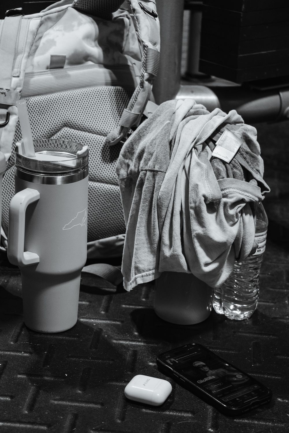 Black and white shot of gym essentials on floor: backpack, water bottle, shirt, tumbler, Airpods, and phone playing music.