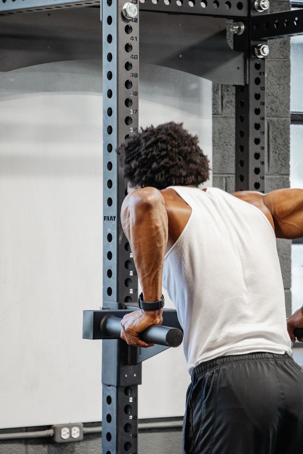Man in white tank top performs dips on parallel bars, arms flexed and focused during upper body workout.
