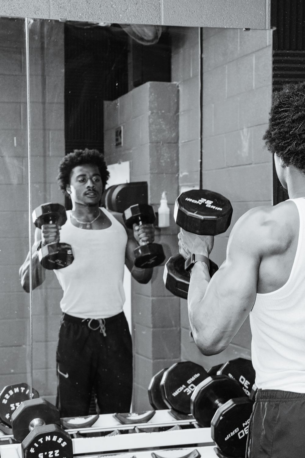 Black & white image of man doing dumbbell curls in front of a gym mirror, muscles flexed and eyes locked on form