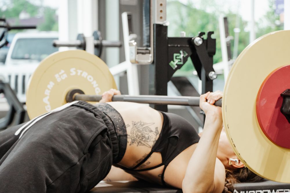 Woman bench pressing with yellow StrongArm plates, tattoo on side.