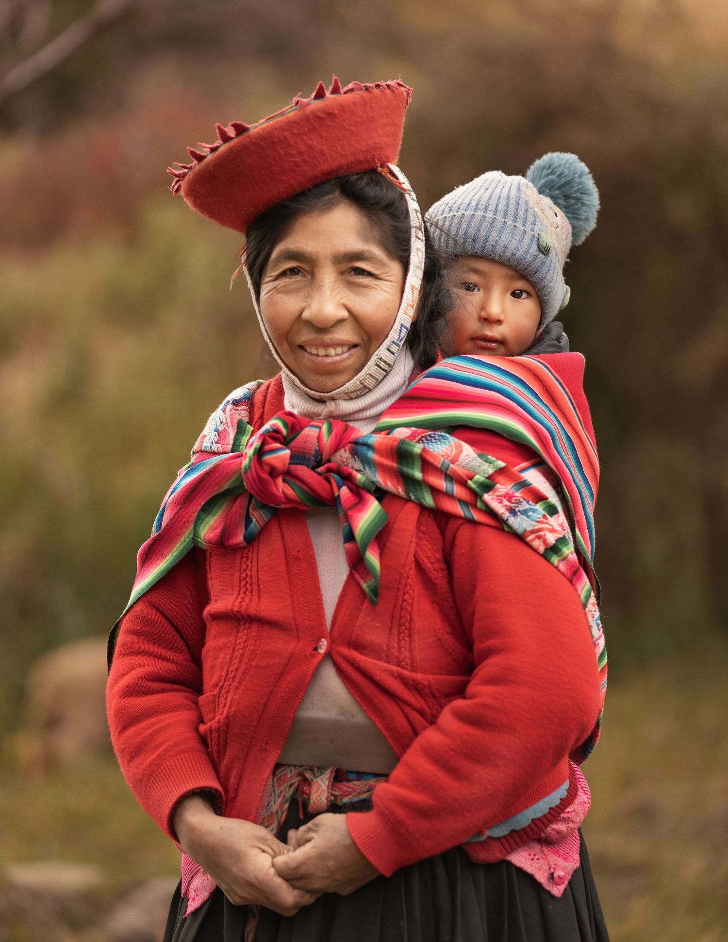 Traditional Peruvian woman in red attire with colorful woven textiles and distinctive hat carries child in traditional style.