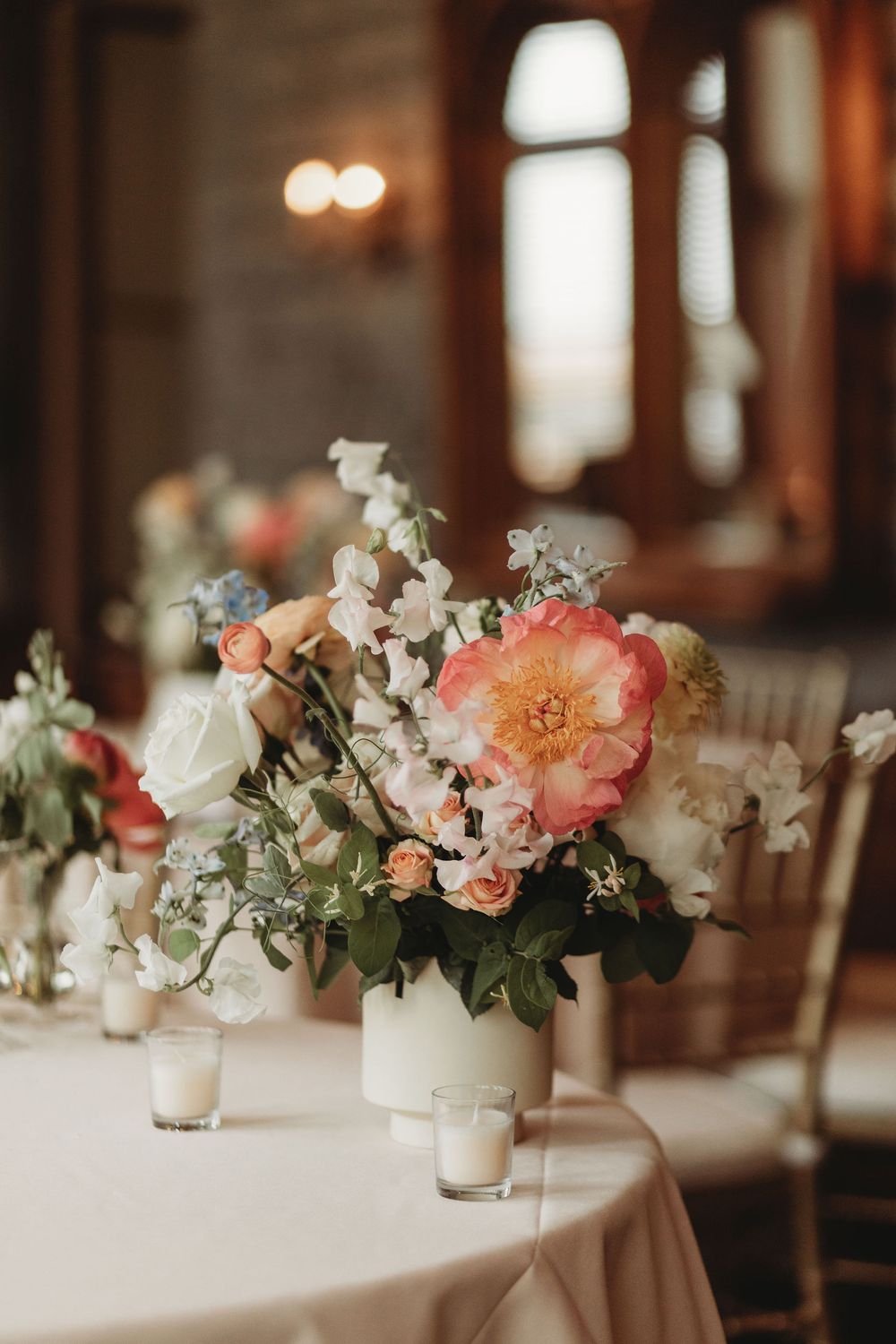 Elegant floral centerpiece with coral peonies and white flowers in white vase on table with candlelight at wedding reception.