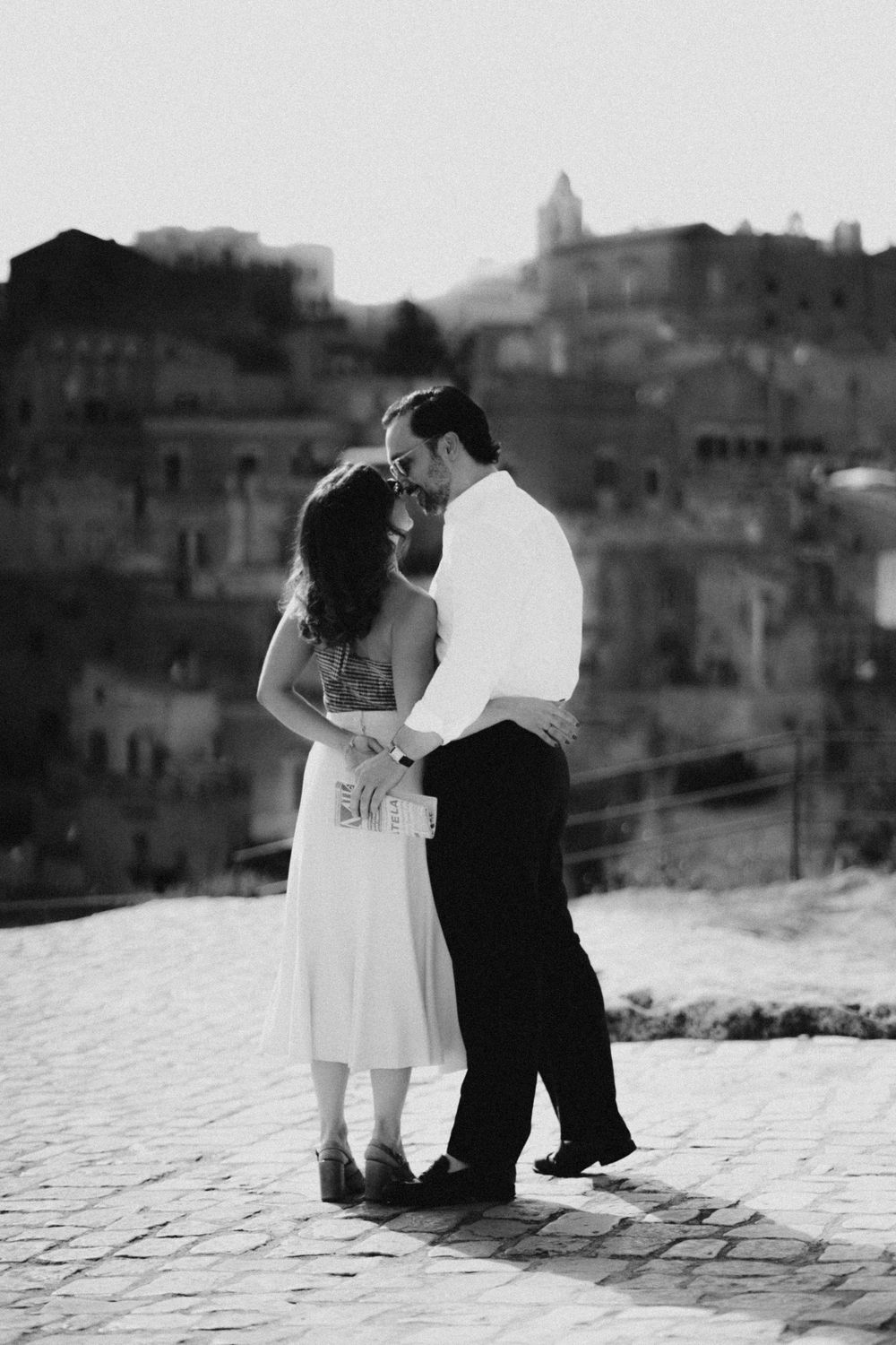 A romantic couple embraces on a cobblestone street overlooking an old Italian hillside town in black and white.