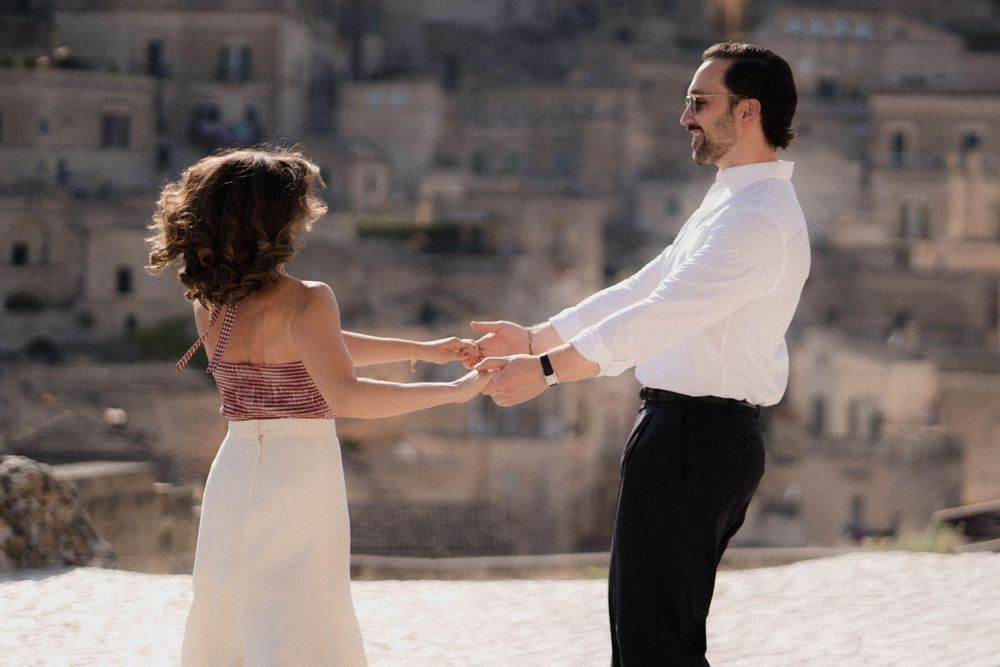A romantic couple holds hands against an ancient stone city backdrop in formal attire during a special occasion.