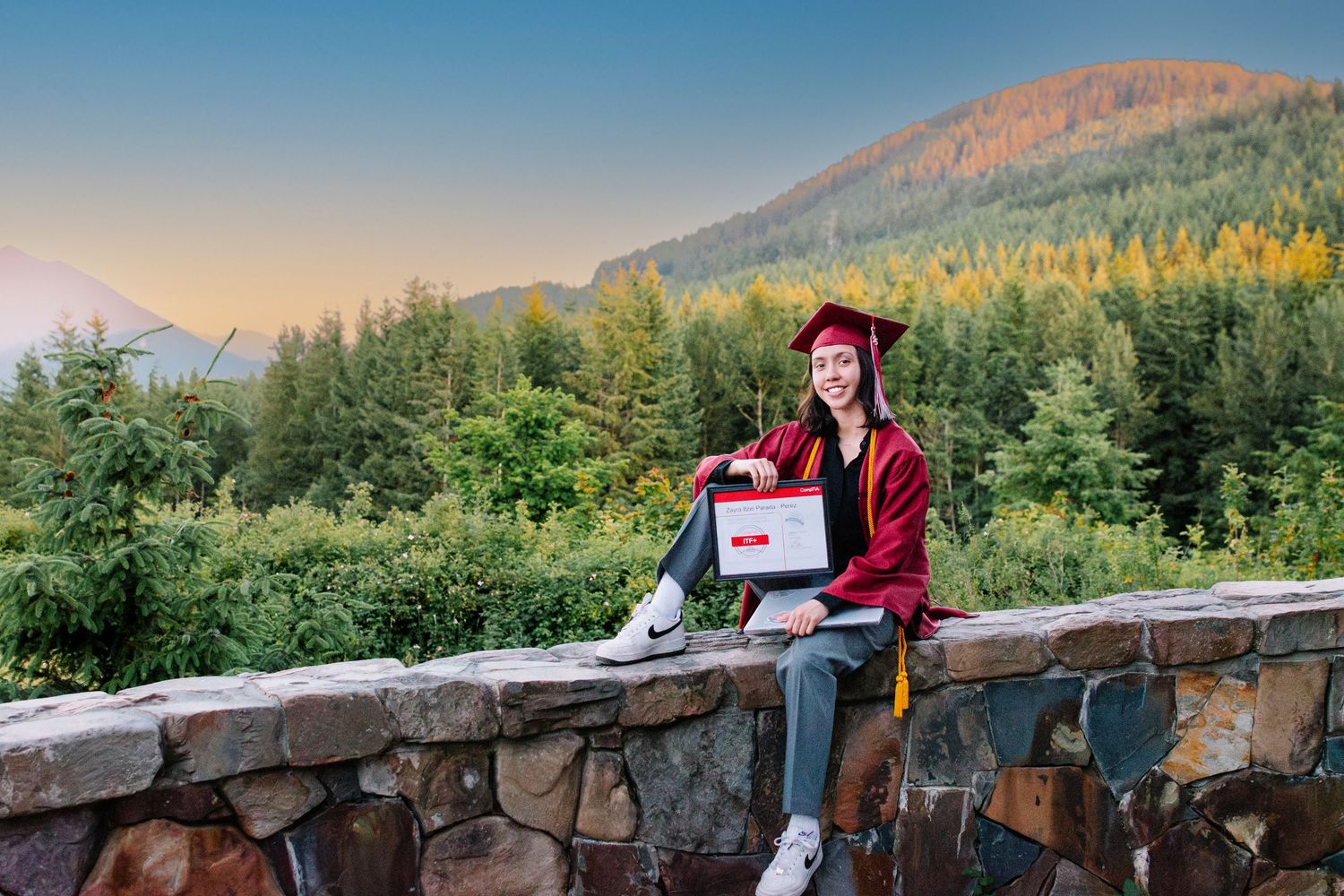 Senior girl seated on stone wall holding diploma case at sunset in the Pacific Northwest. Snoqualmie Point Park. Seattle Graduation Photo.